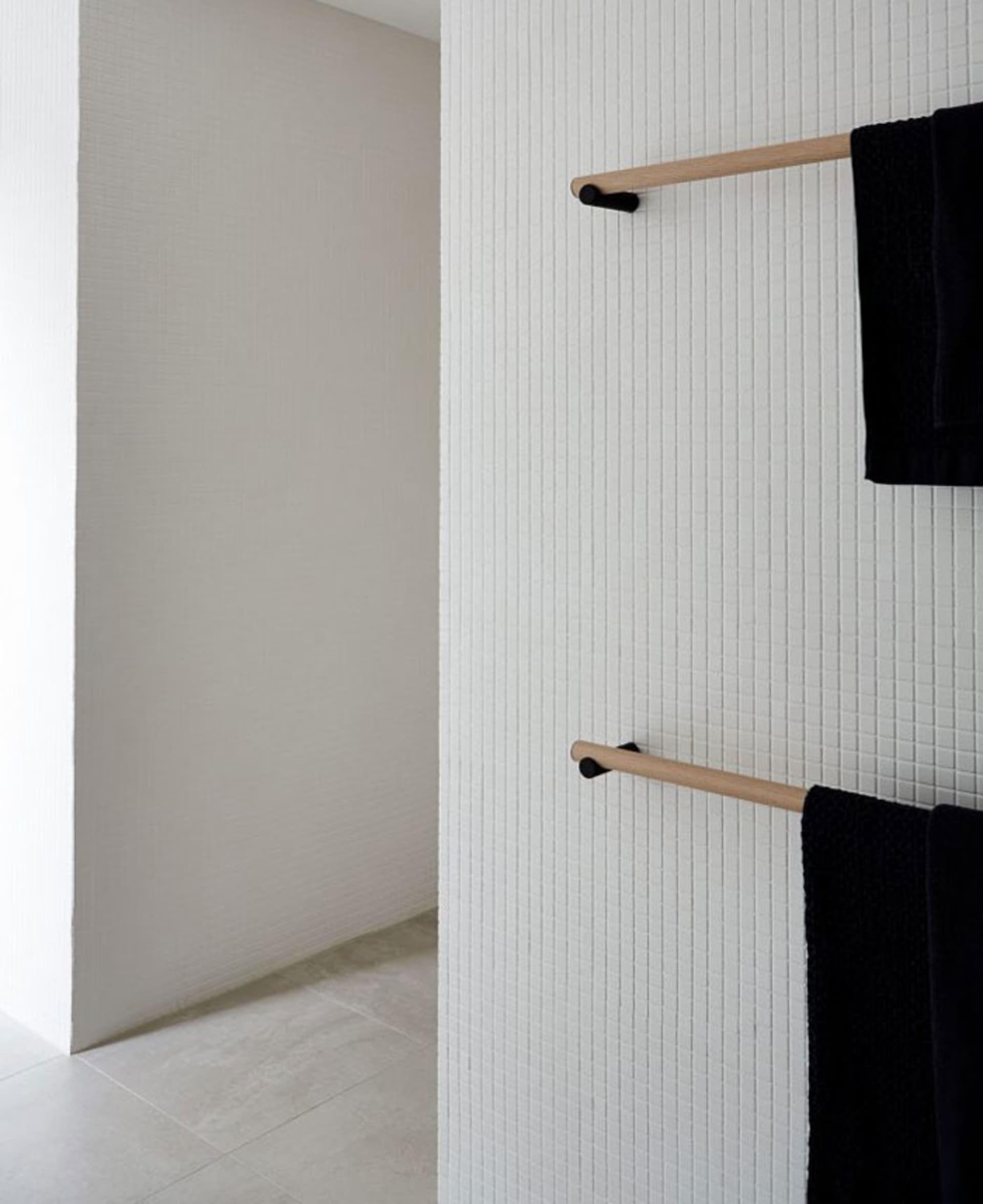 A modern minimalist bathroom with white ceramic tiles and black towels hanging on a wooden rack.