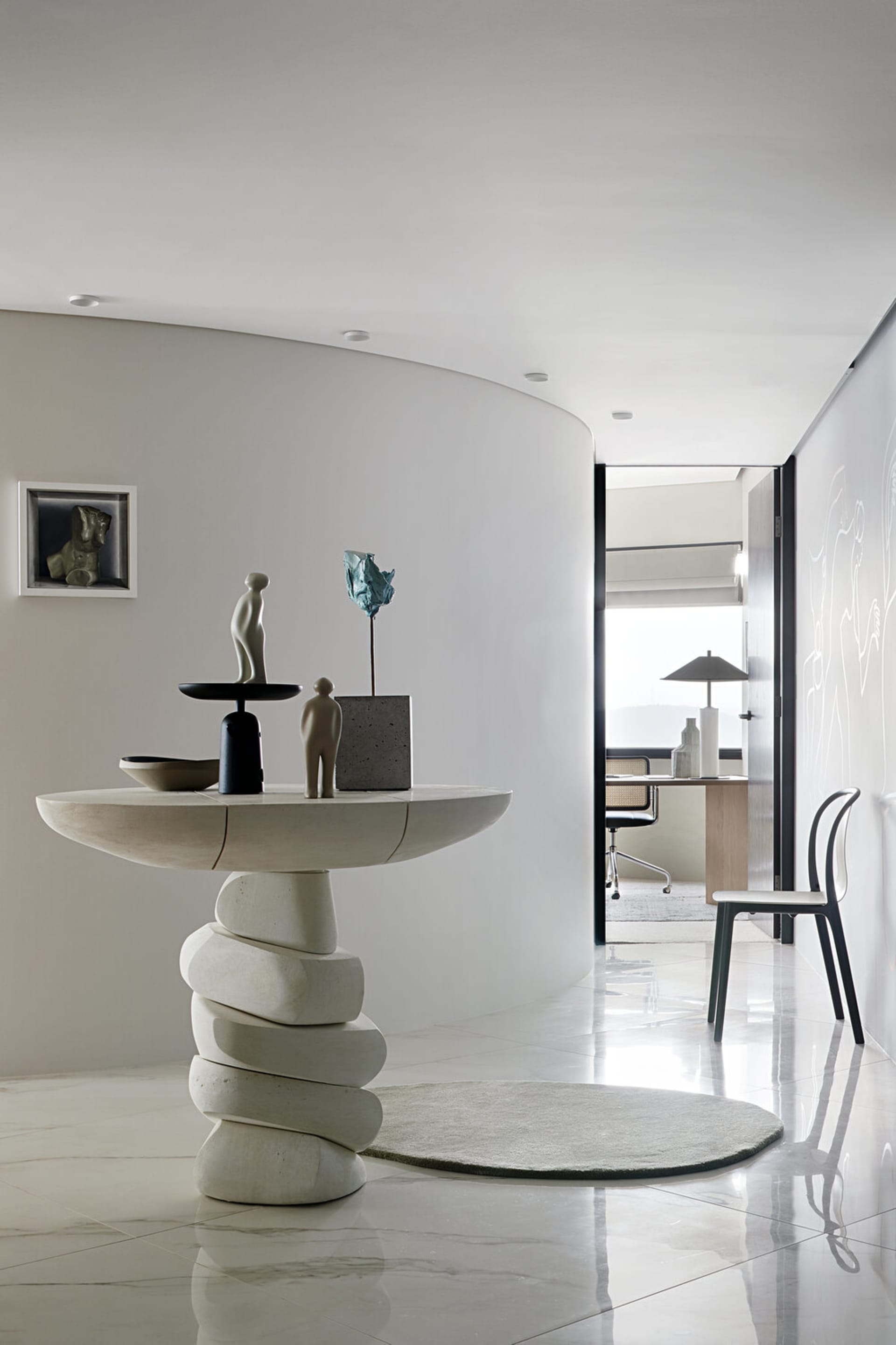 A modern minimalist entryway featuring a concrete table with various sculptures on top, set against a white wall and floor.