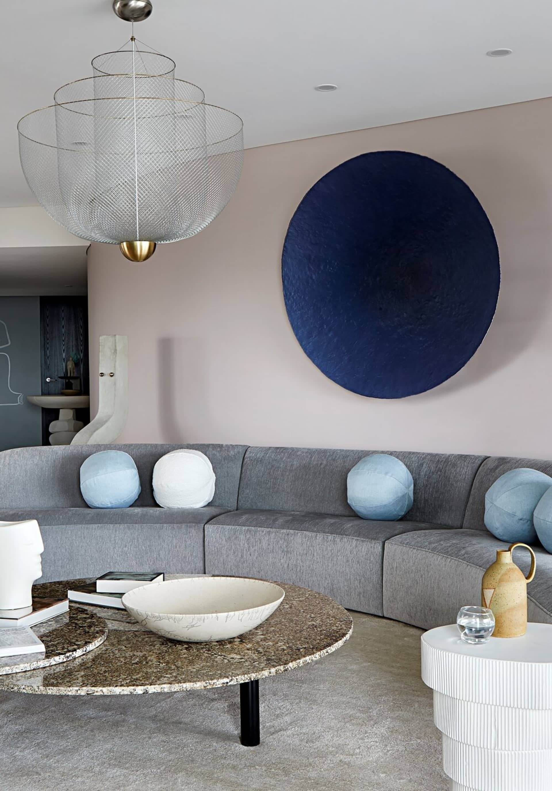 A modern minimalist living room with a curved gray sofa adorned with light blue cushions, a round coffee table with a stone t