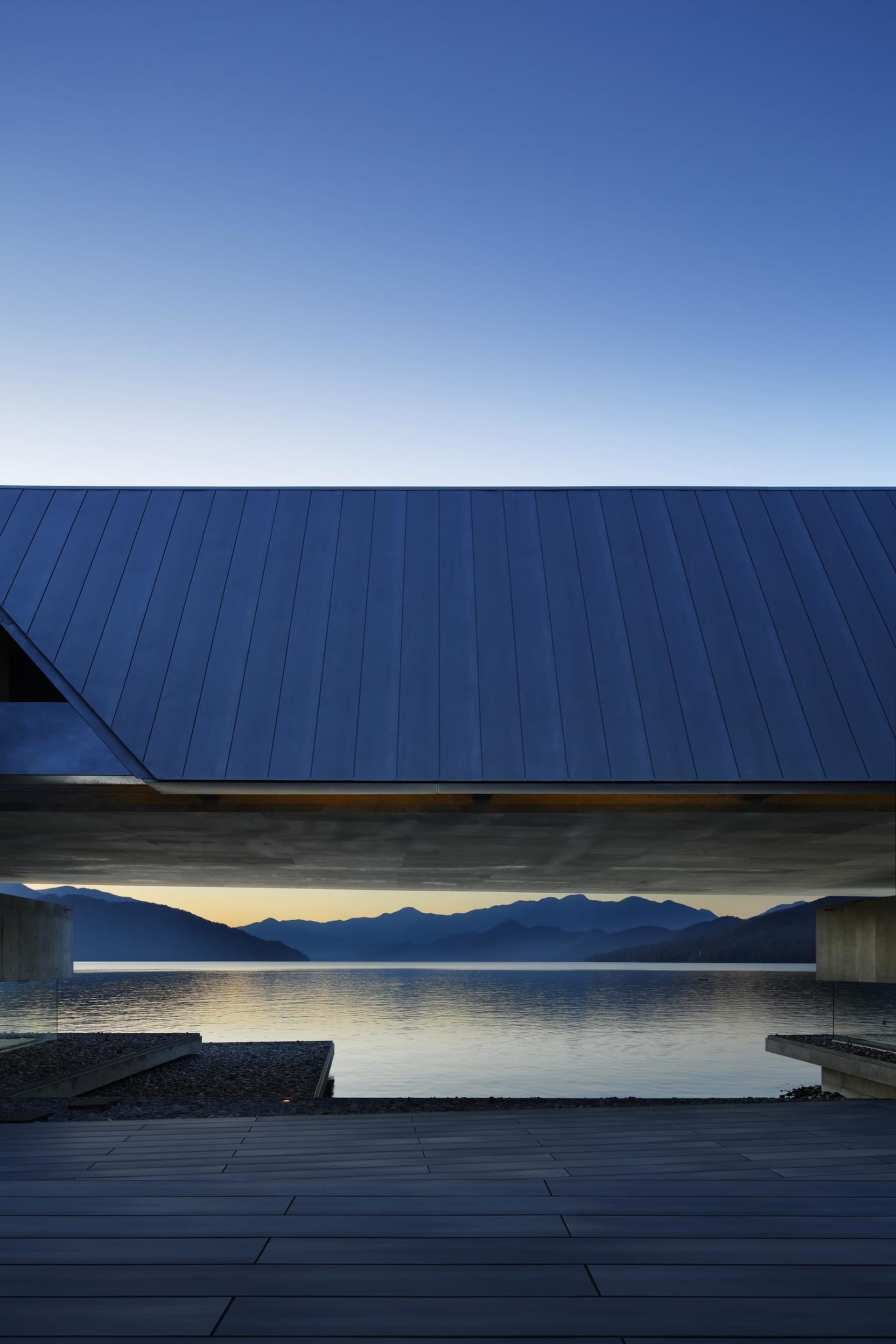 A modern structure with a metal roof and concrete floors overlooking a lake and distant mountains.