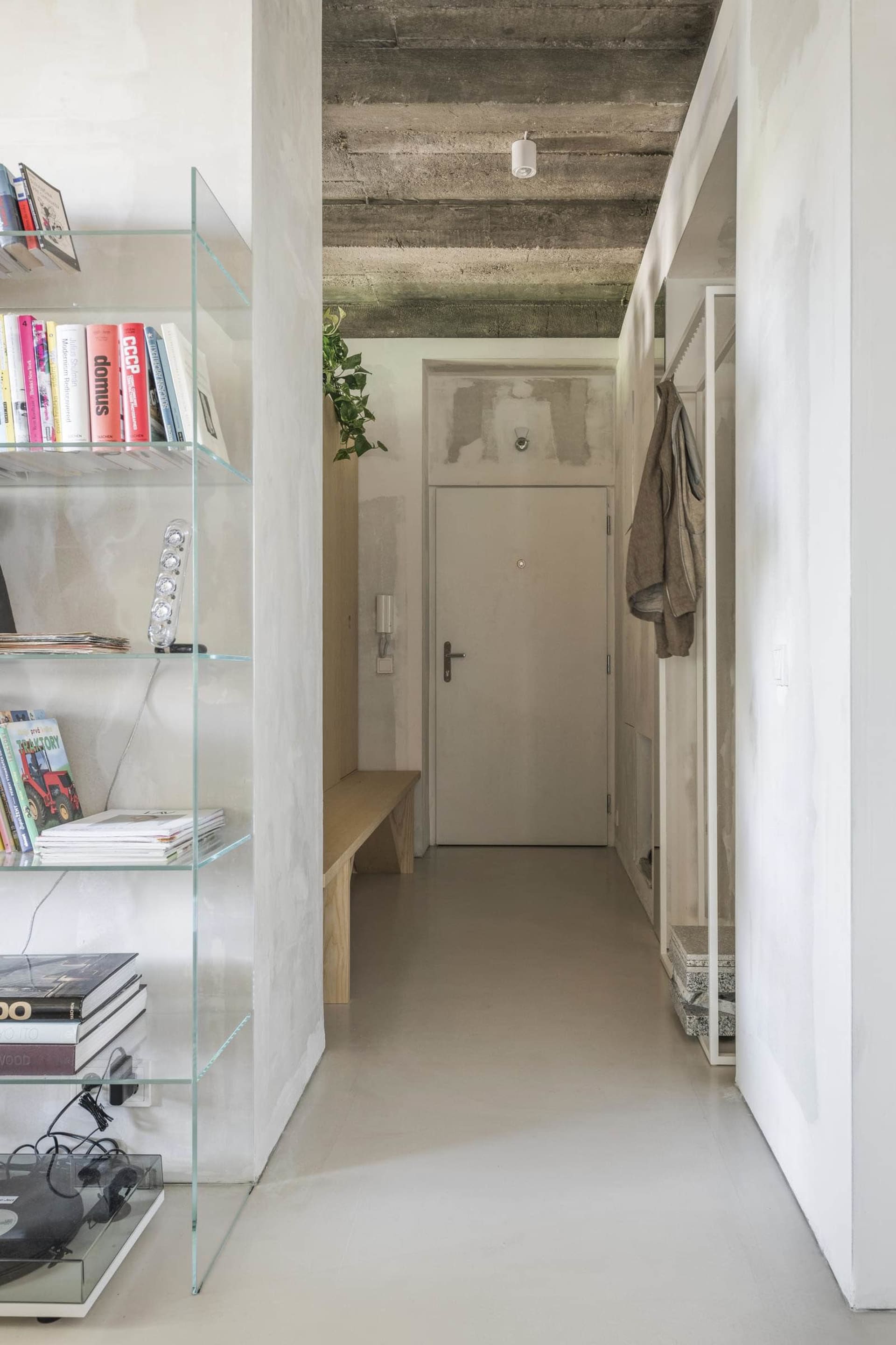 Modern minimalist entryway with wooden bench, glass bookshelf, and concrete ceiling.
