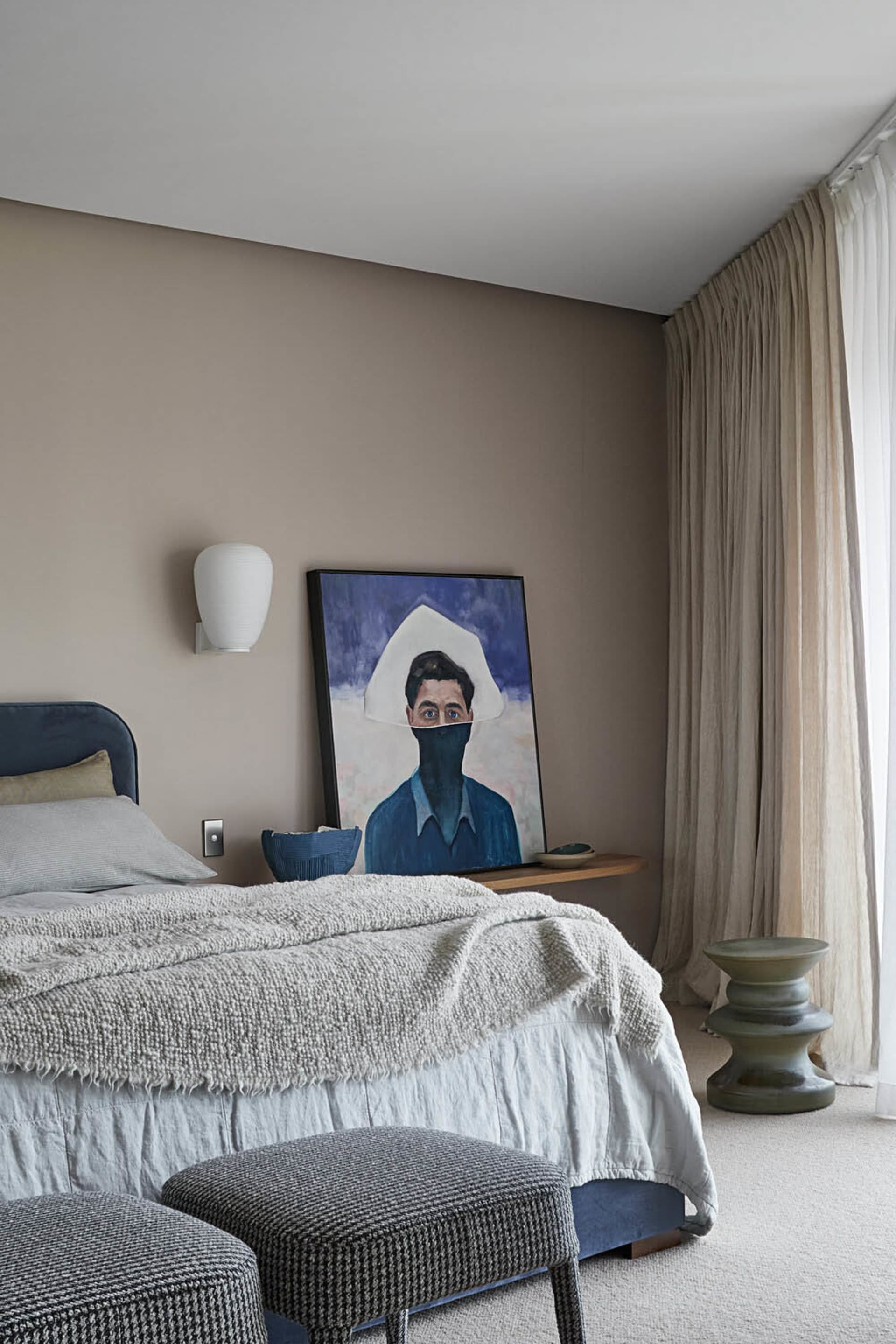 A modern bedroom featuring a bed with gray bedding, a dark blue bed frame, and a hanging sconce above it, alongside a portrai