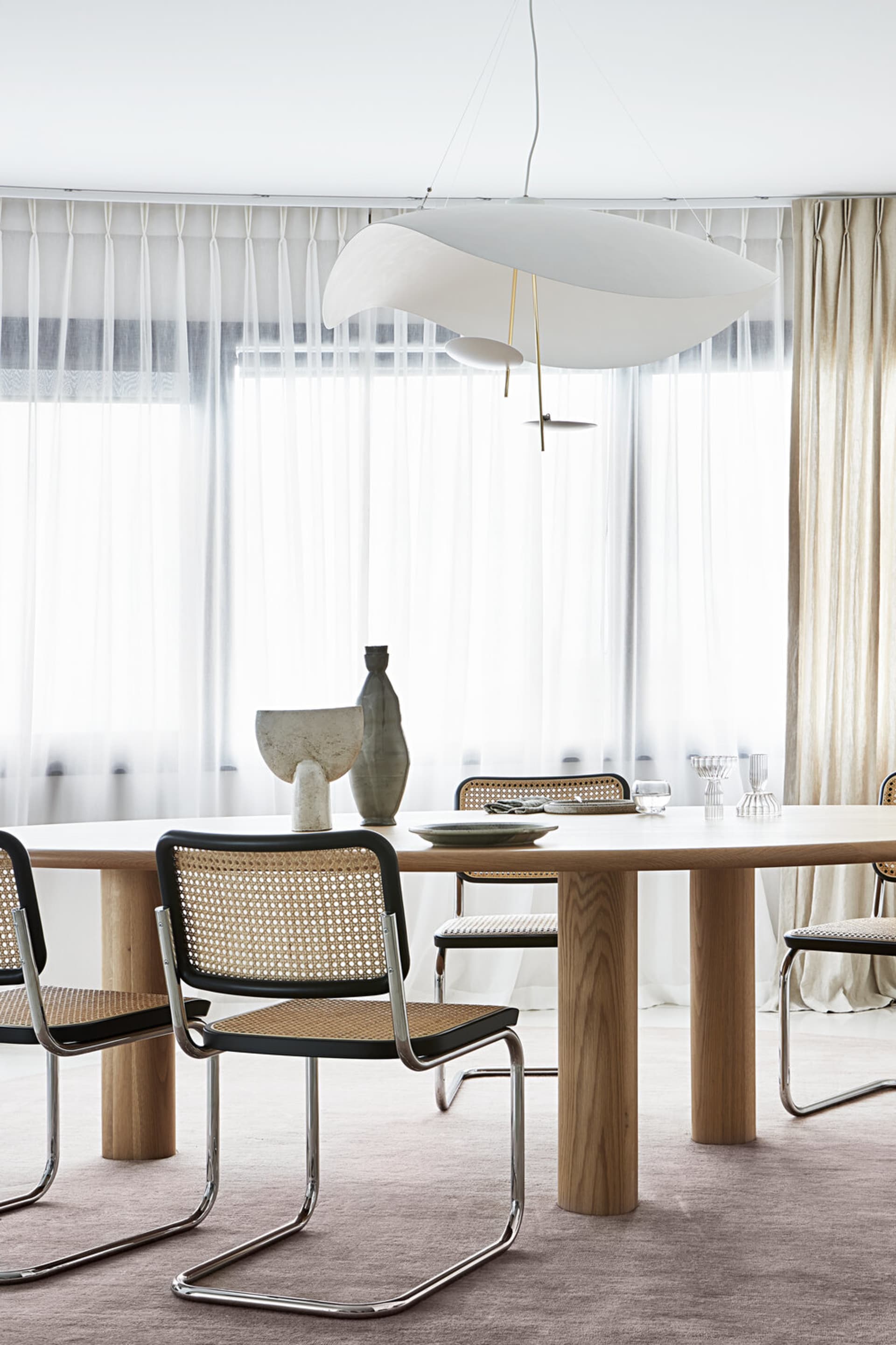 A modern dining room with a wooden table, black and rattan chairs, and a large window with sheer curtains.