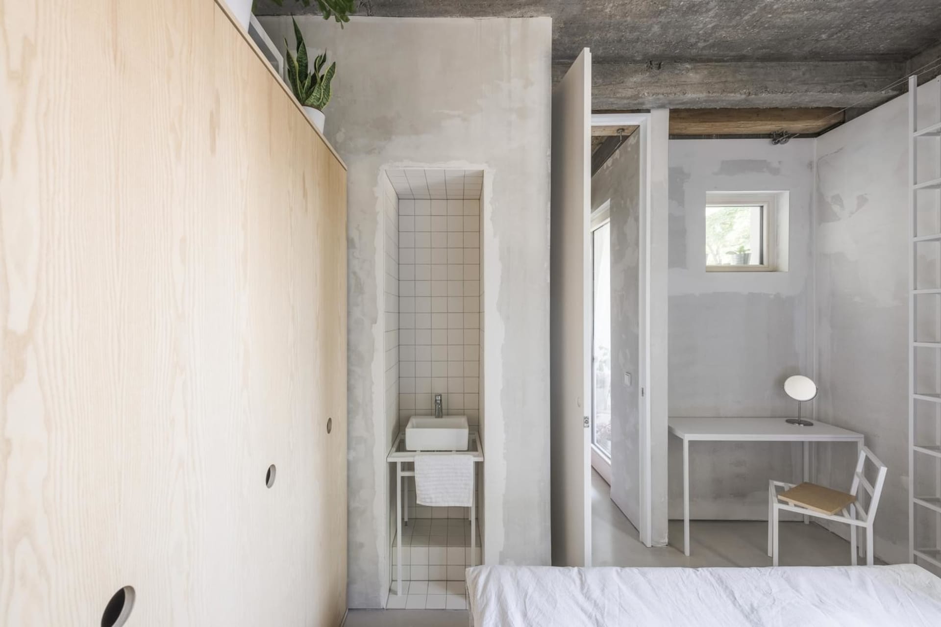 A modern minimalist bedroom with raw concrete walls and a white bed, featuring a small bathroom area with a tiled floor and s
