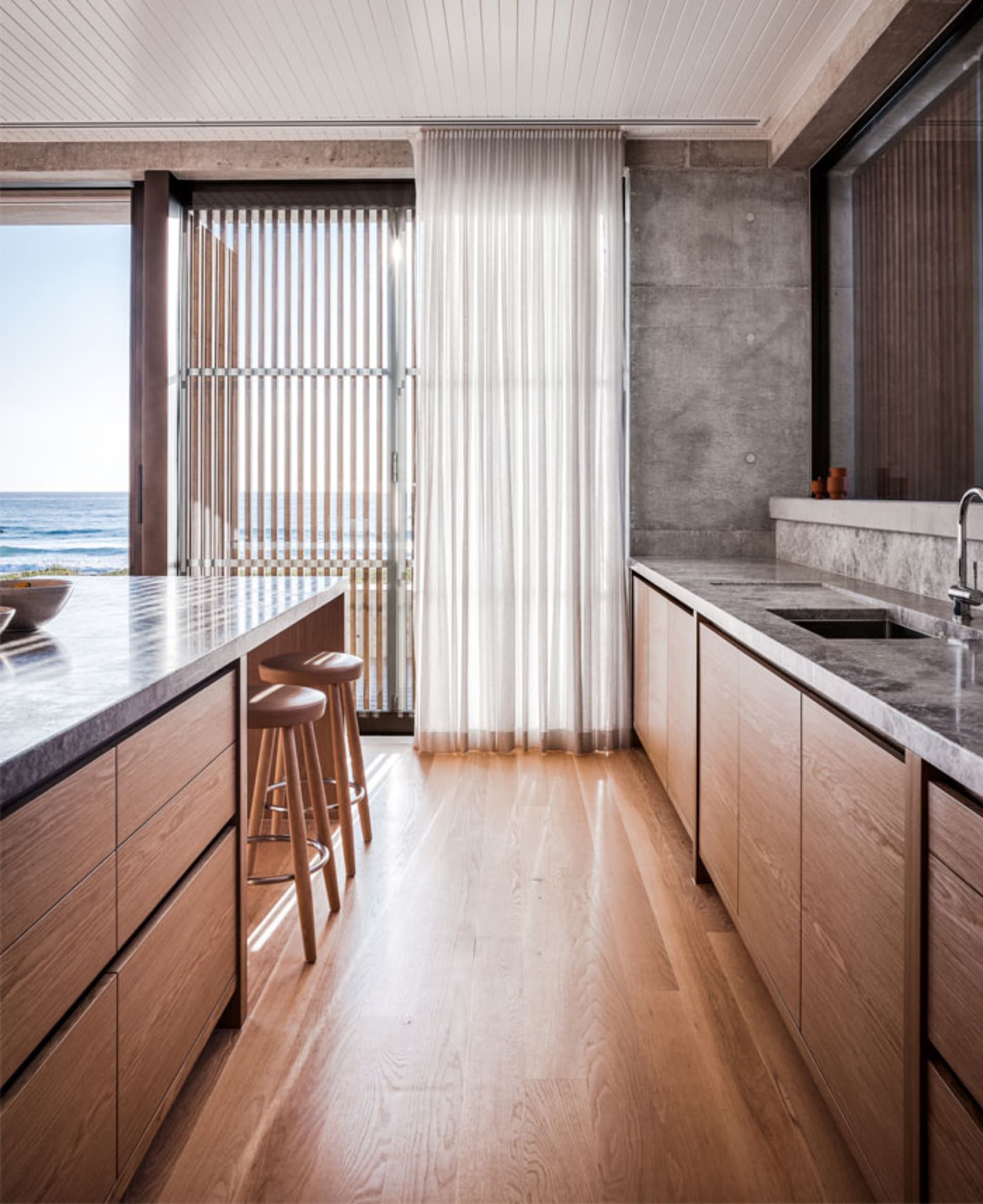 Modern kitchen with wooden cabinets, marble countertops, and a view of the ocean through large windows with vertical blinds a