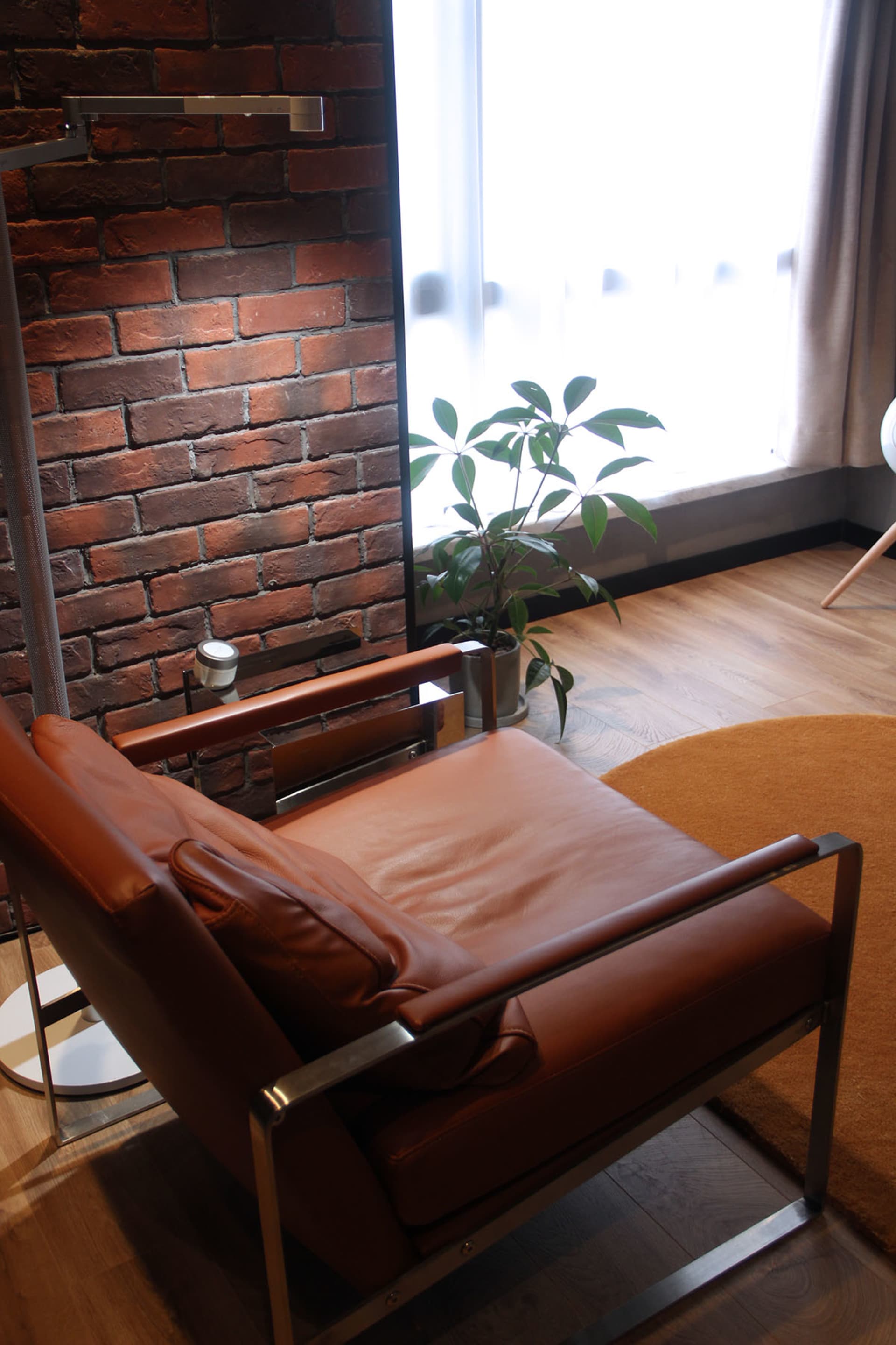 A cozy industrial-style living room features a modern leather chair, metallic frame, wooden floor, and brick accent wall.