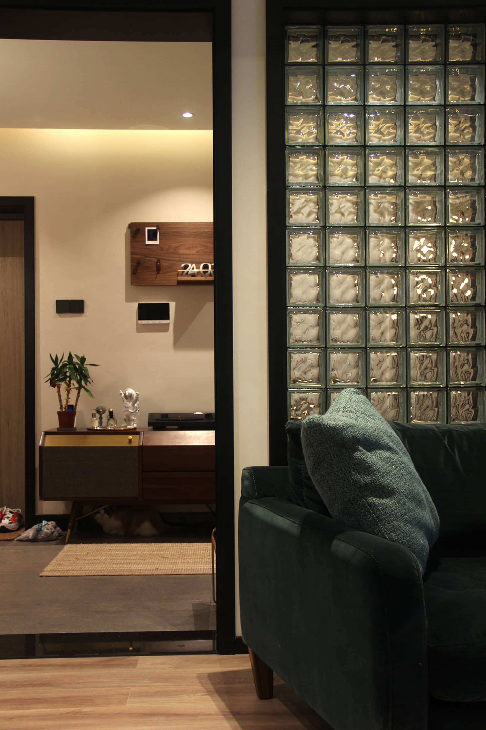 A modern living room featuring a glass block wall, a green velvet sofa, and a wooden cabinet with a potted plant and a small