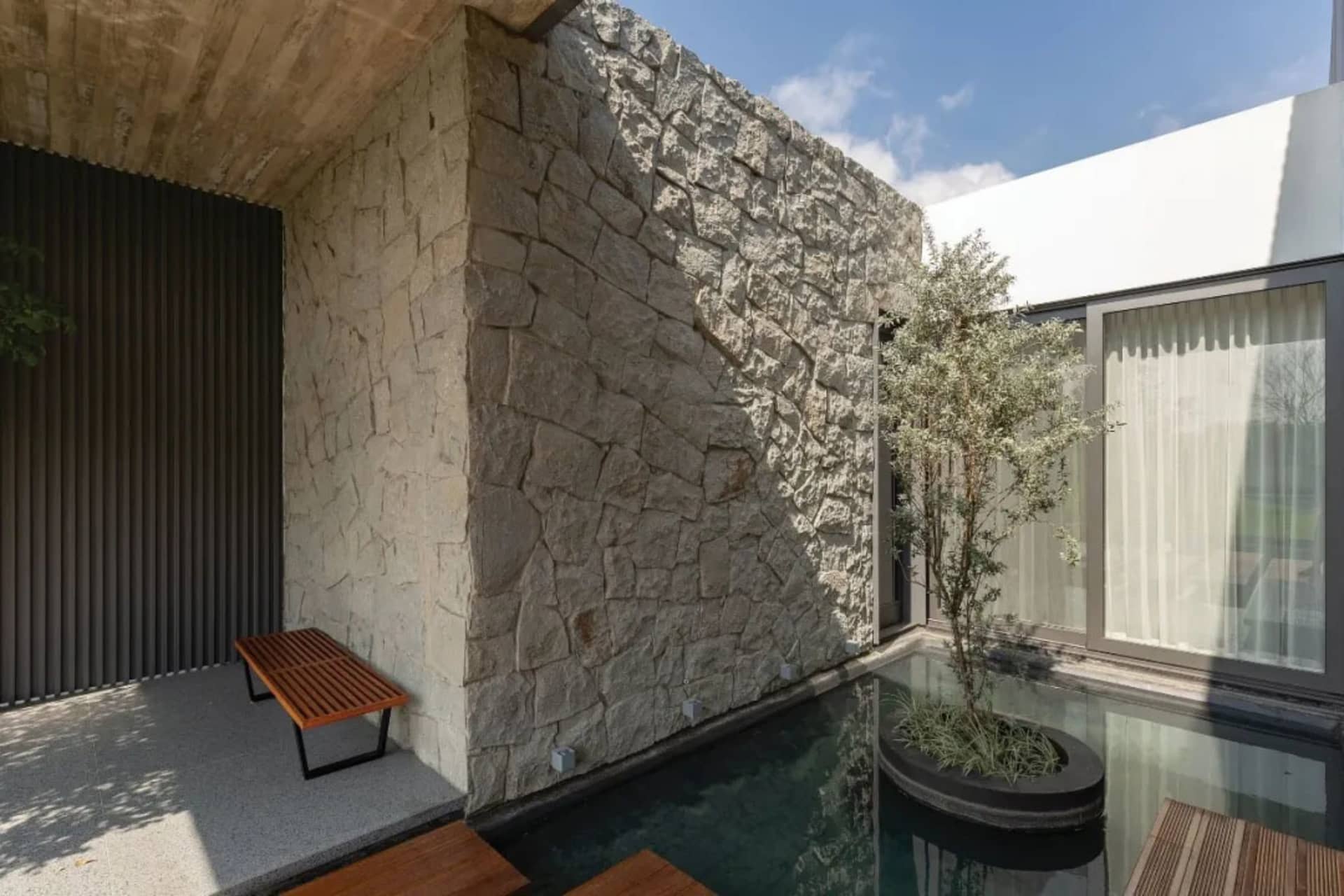 A modern minimalist residential space featuring a concrete and stone wall, a glass poolside area, and a wooden bench.