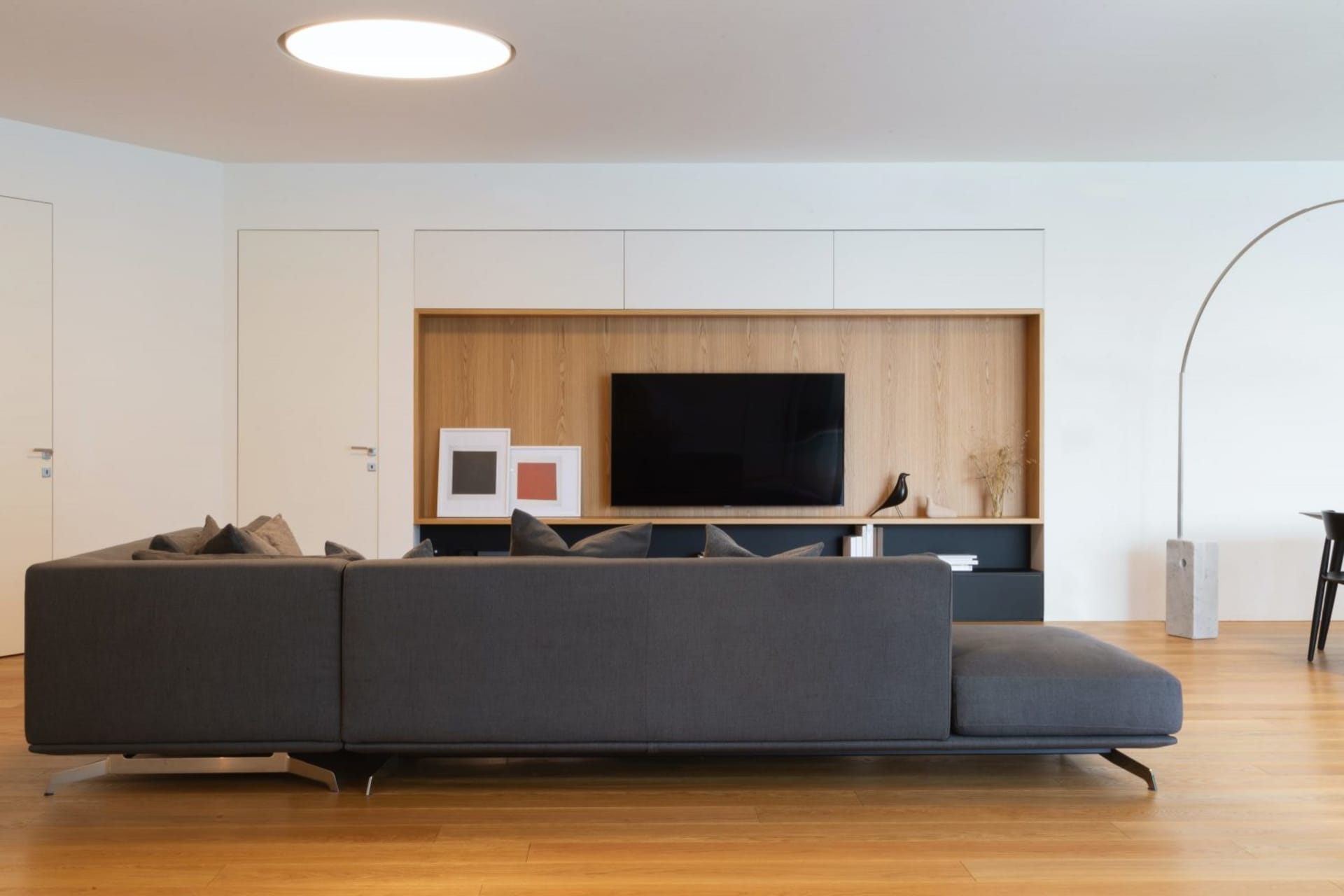 Modern living room with a dark gray sofa, wooden furniture, and a television mounted on a wooden panel with framed artwork.
