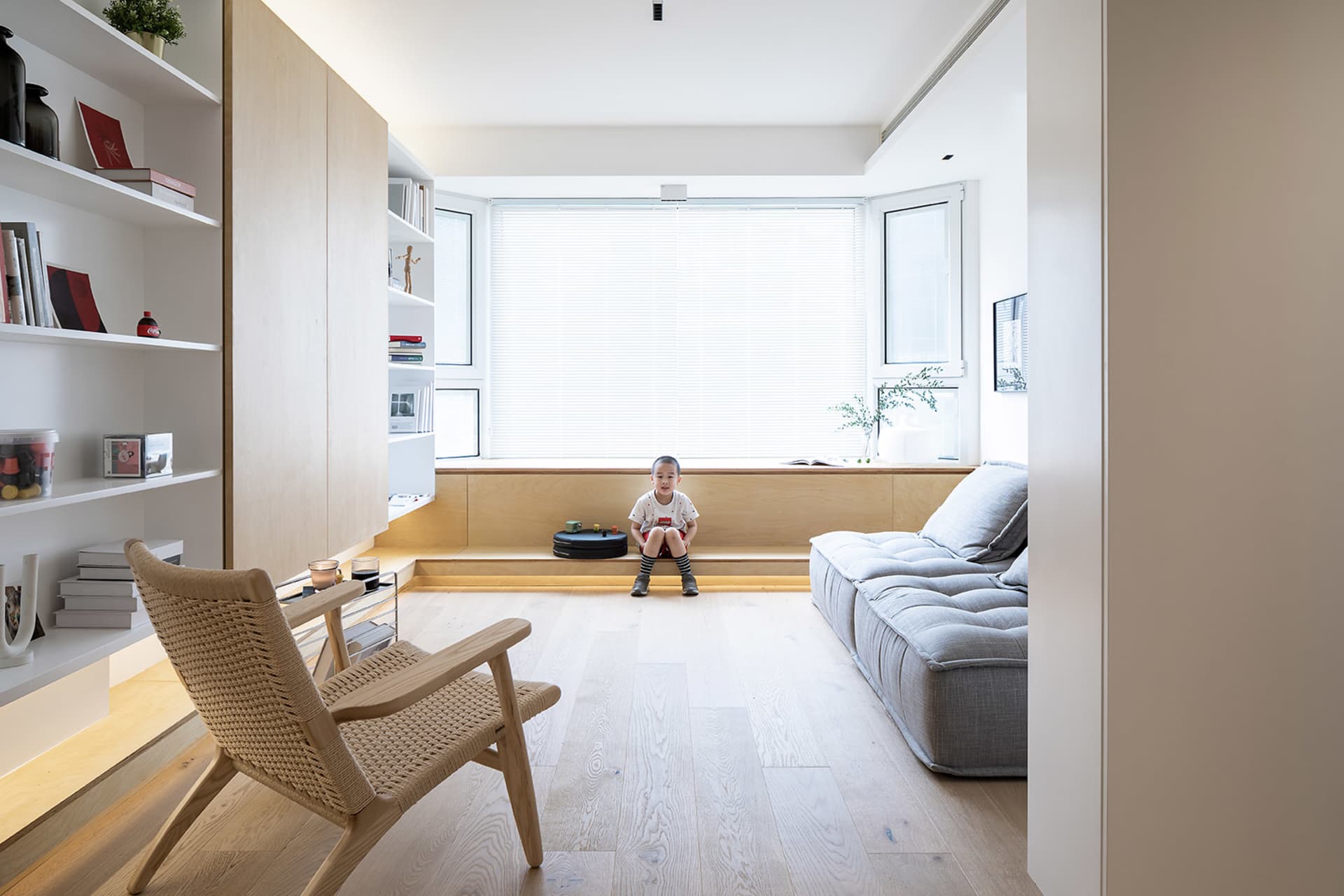 A minimalist family living room with a child sitting on a window seat, a grey sofa, a woven armchair, and built-in shelves