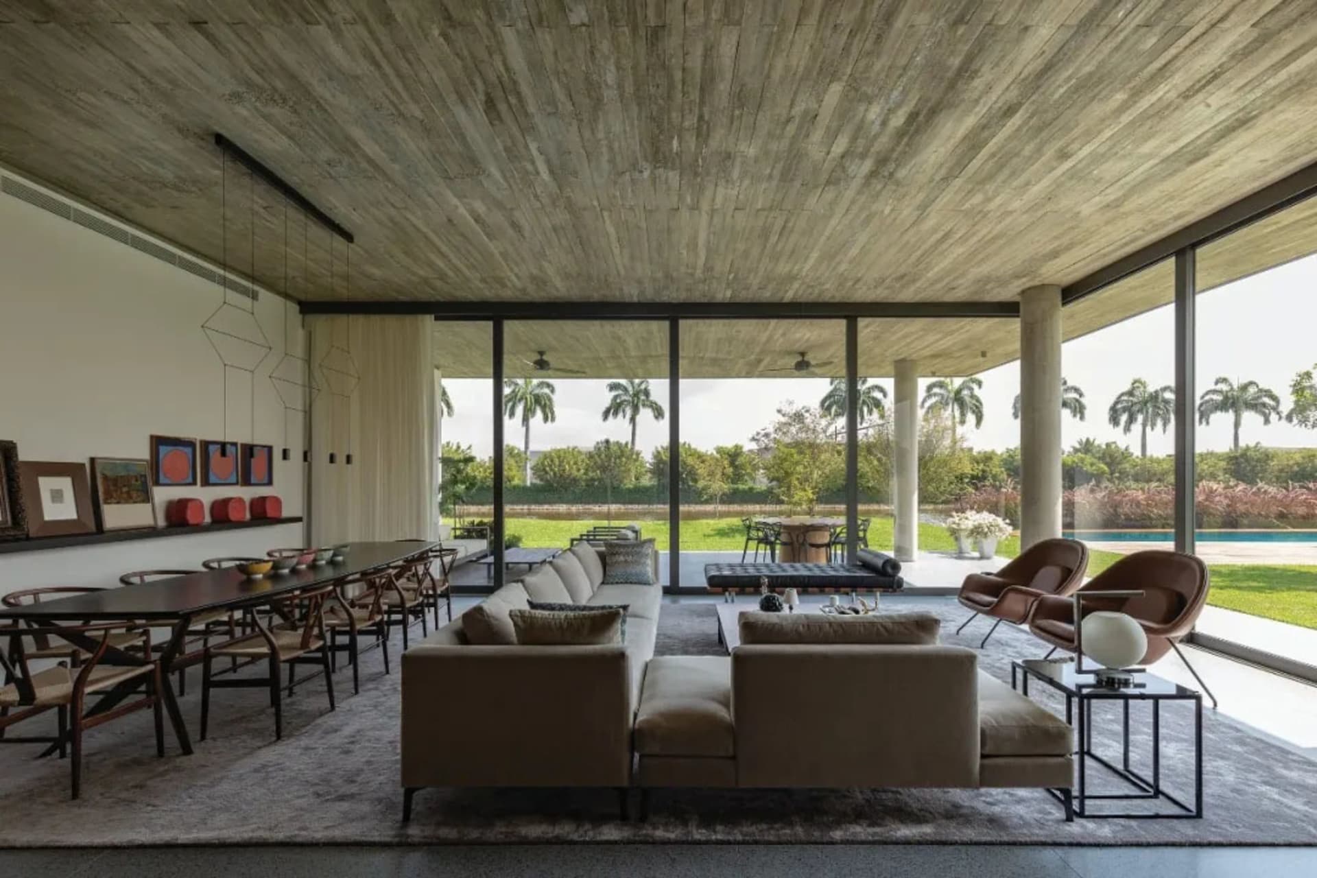 Modern living room with concrete walls, large windows overlooking a pool and garden, and a comfortable sofa and dining table.