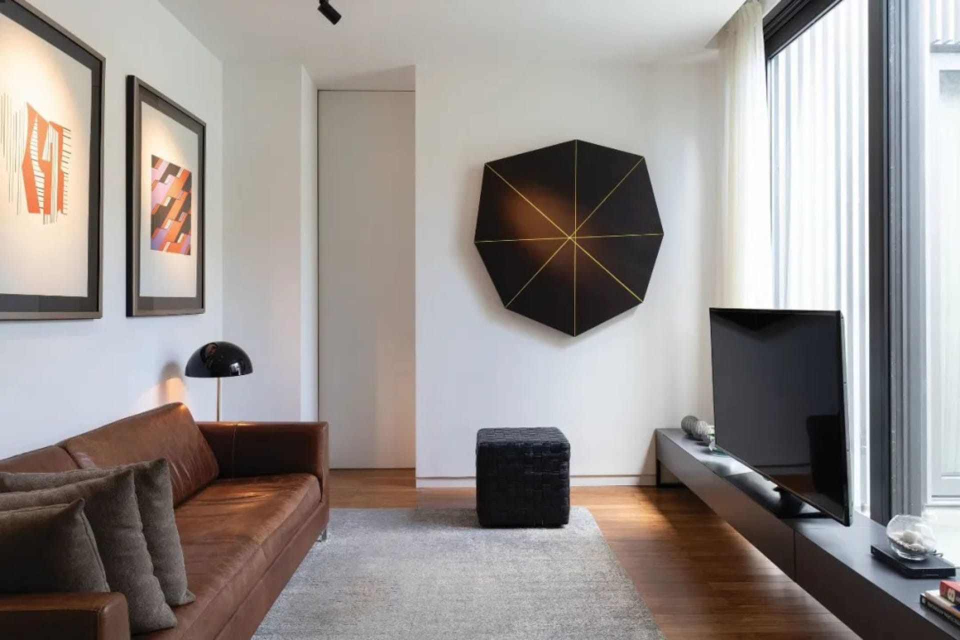 A modern living room featuring a brown leather sofa, a black geometric art piece on the wall, and a television on a floating