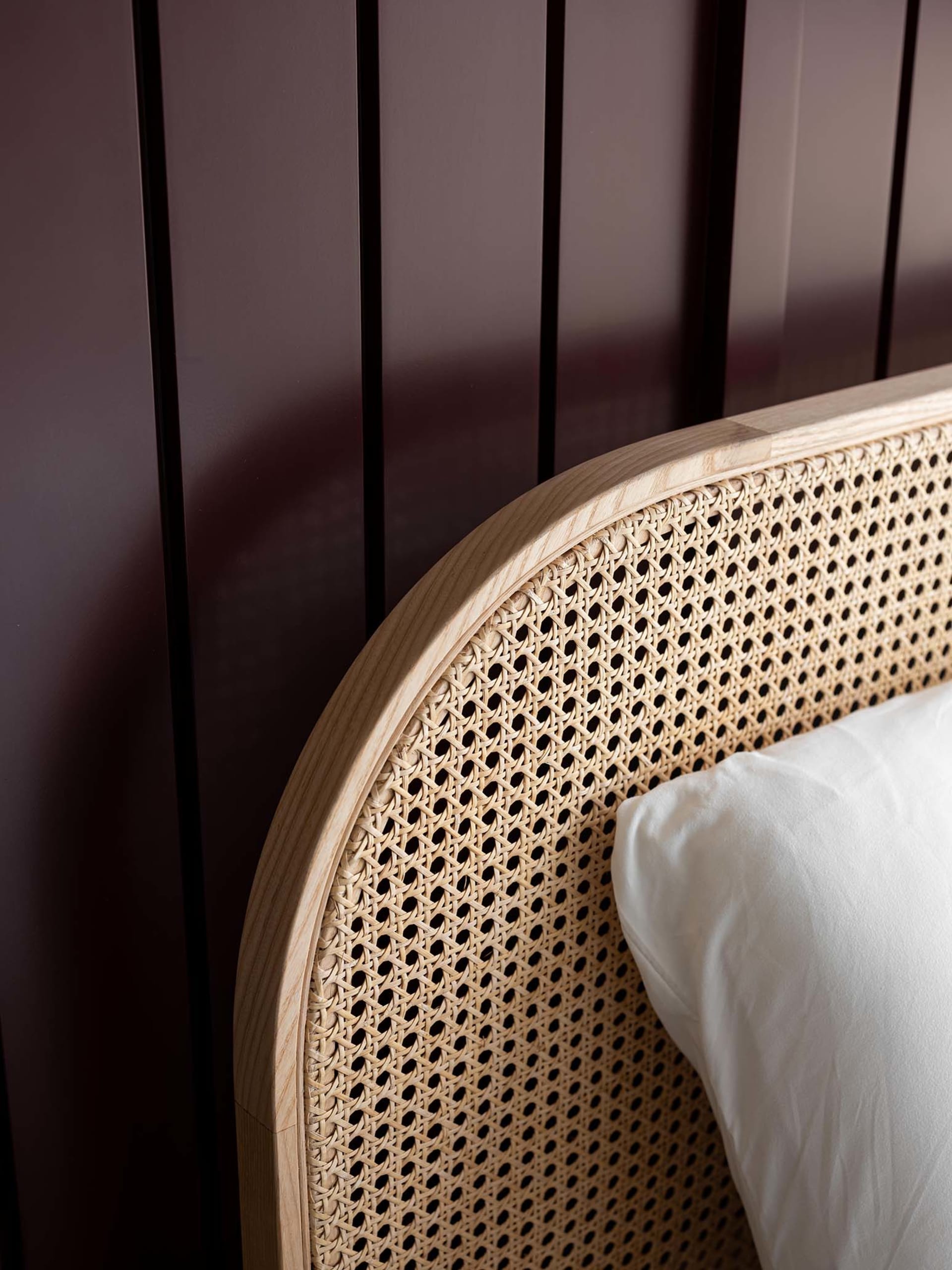 A modern bedframe with a woven cane headboard and a white pillow against a dark brown wooden wall.