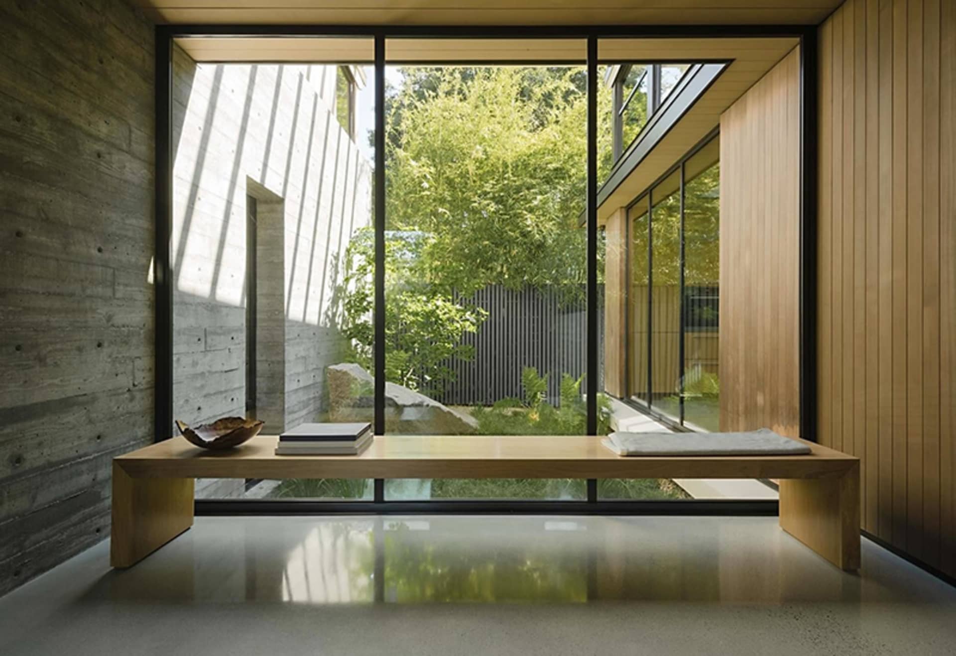 A modern interior space featuring wooden furniture, concrete walls, and large windows overlooking a garden.