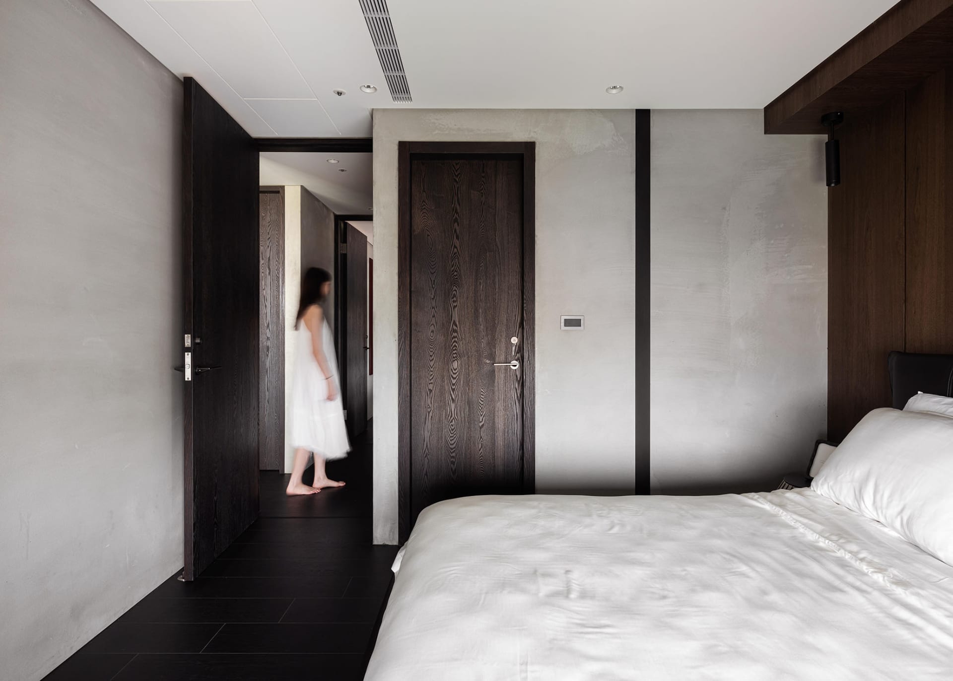 A minimalist bedroom with concrete walls, a white bed, and a person walking through the hallway.