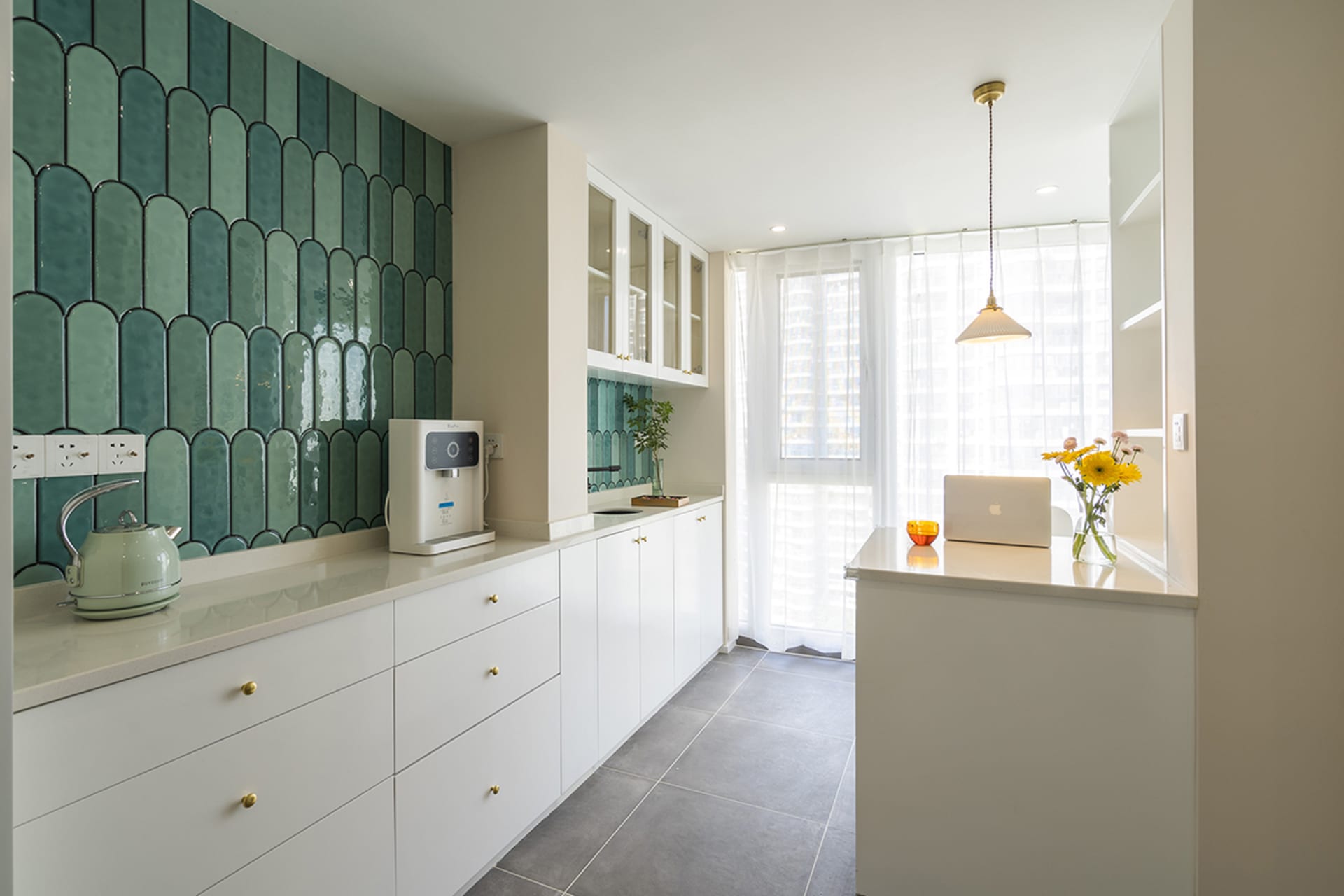 A modern, minimalist kitchen featuring green ceramic tiles, white cabinetry, and a white countertop with a kettle, laptop, an