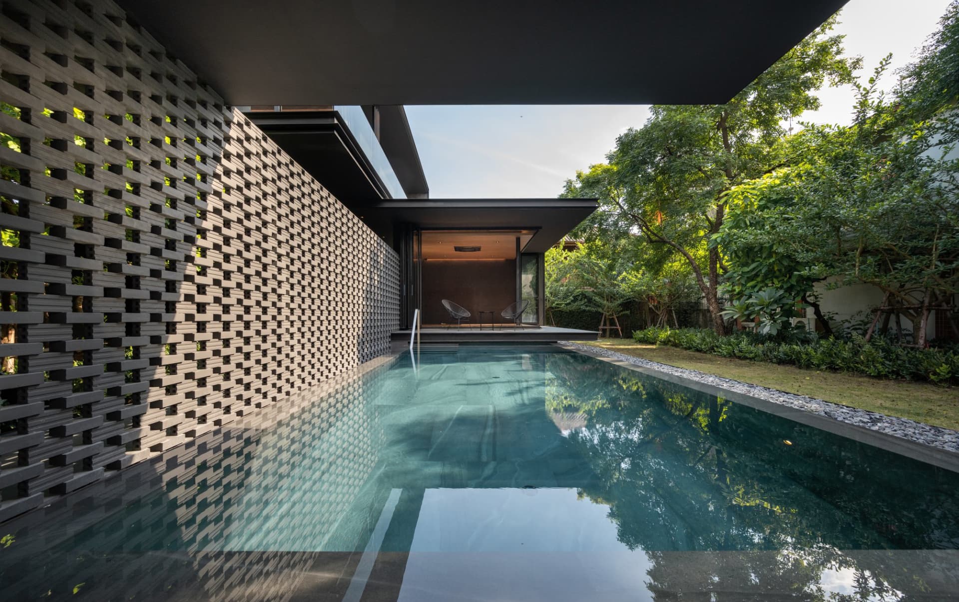 A modern residential exterior featuring a concrete pool surrounded by greenery and a black lattice wall.