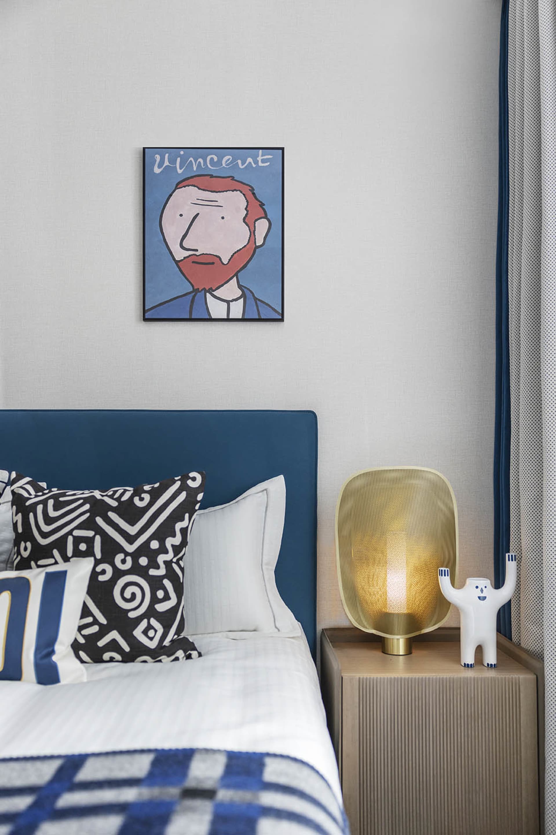 A modern bedroom featuring a blue upholstered headboard with patterned throw pillows and a golden lamp on a side table.