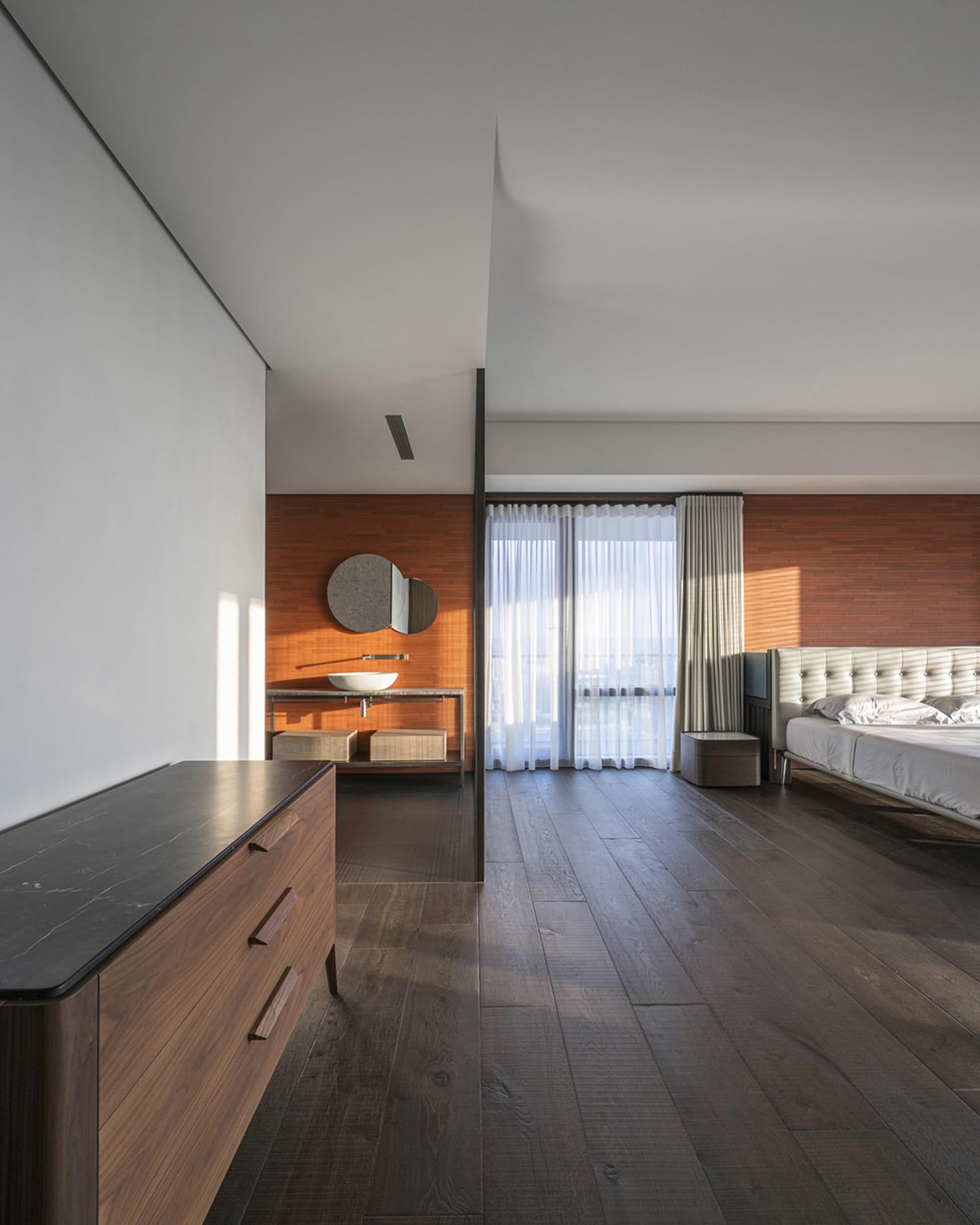 A modern bedroom with a wooden floor, a white bed, and a sink in the background.