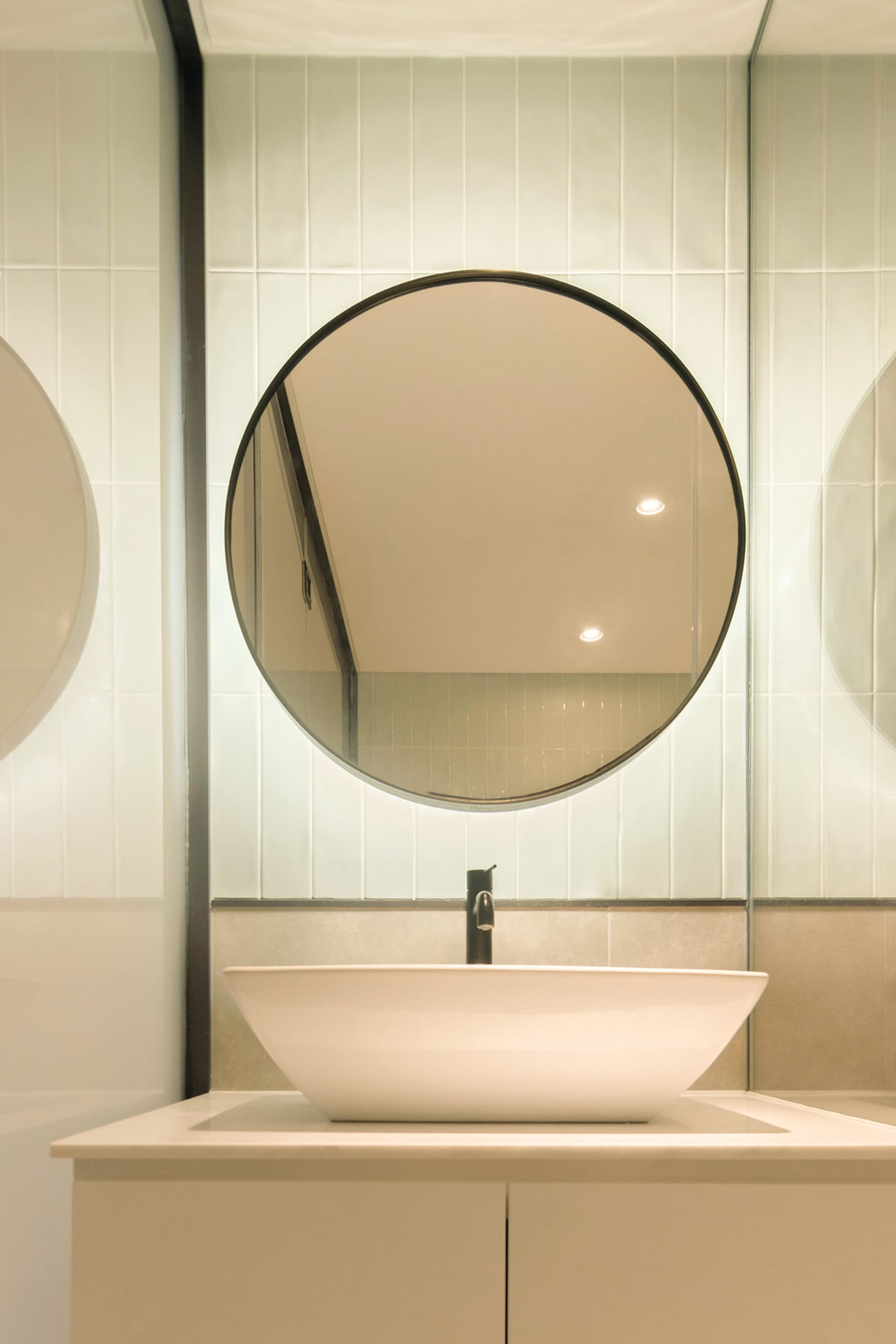 A modern bathroom features a circular mirror hanging above a white sink, surrounded by ceramic tiles with a sleek, minimalist