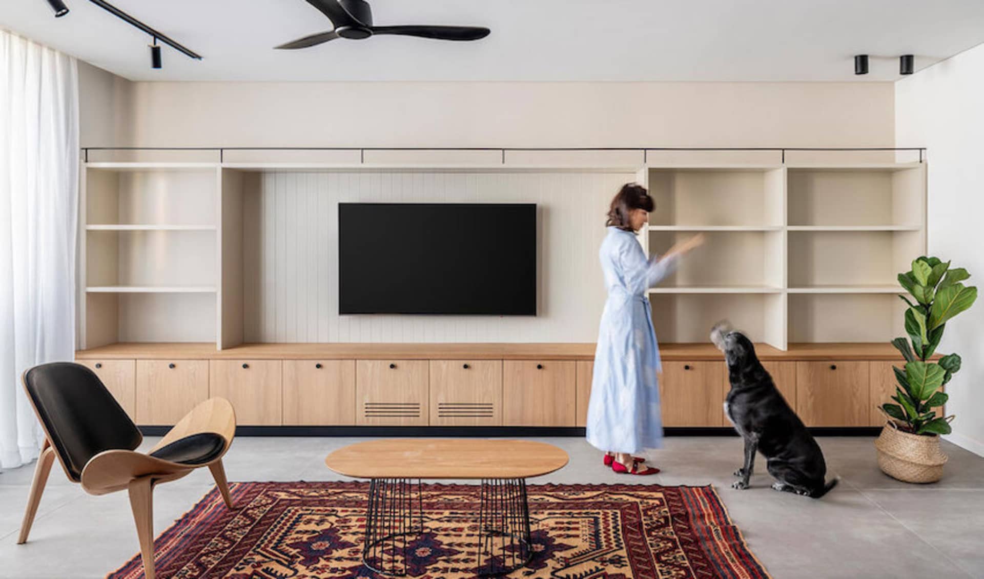 A modern living room with a wooden coffee table, a black armchair, and a woman arranging items on a white shelving unit.