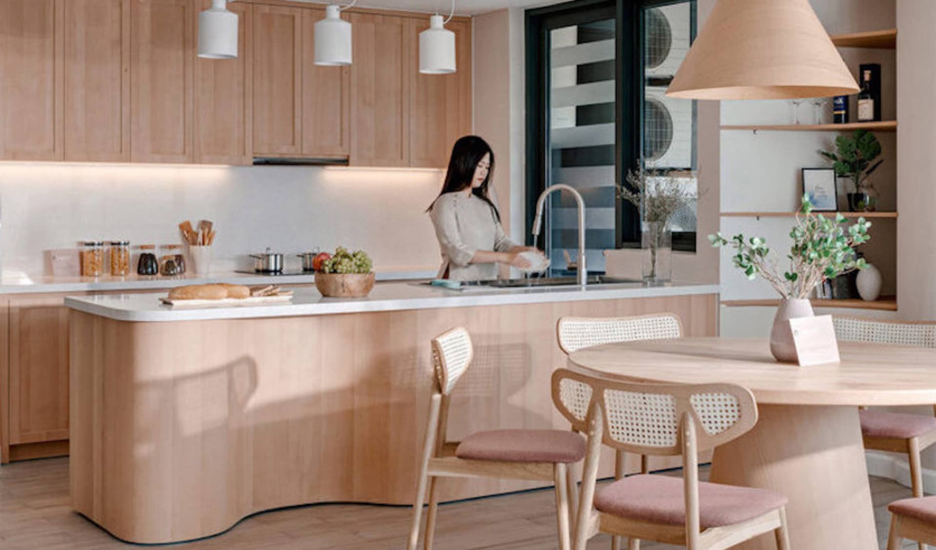 A modern kitchen with light-colored wood cabinets and a matching breakfast bar, featuring a woman washing dishes at the sink.