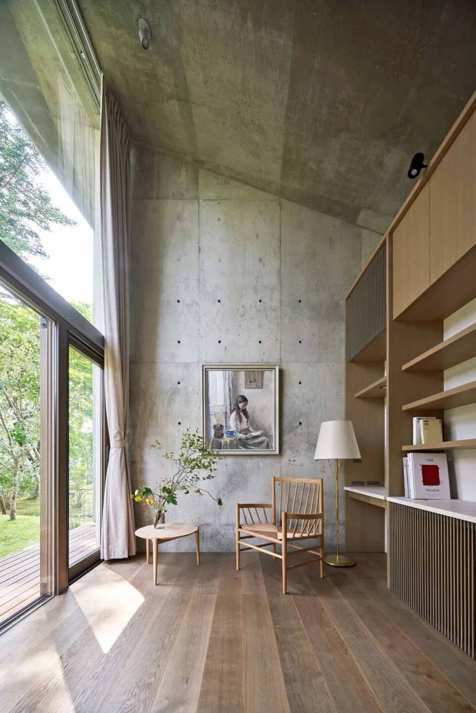 A modern minimalist living room featuring concrete walls, wooden furniture, and large windows with a garden view.