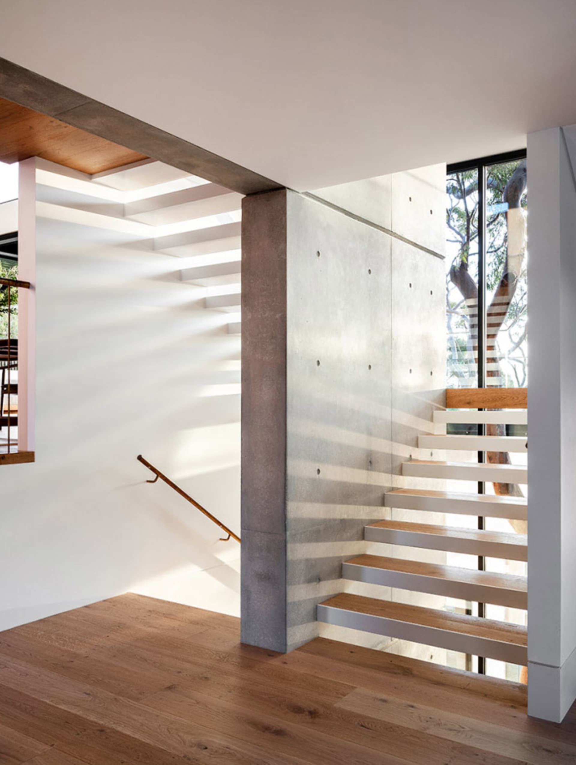 Modern architectural interior with concrete walls, wooden stairs, and large windows showing trees.