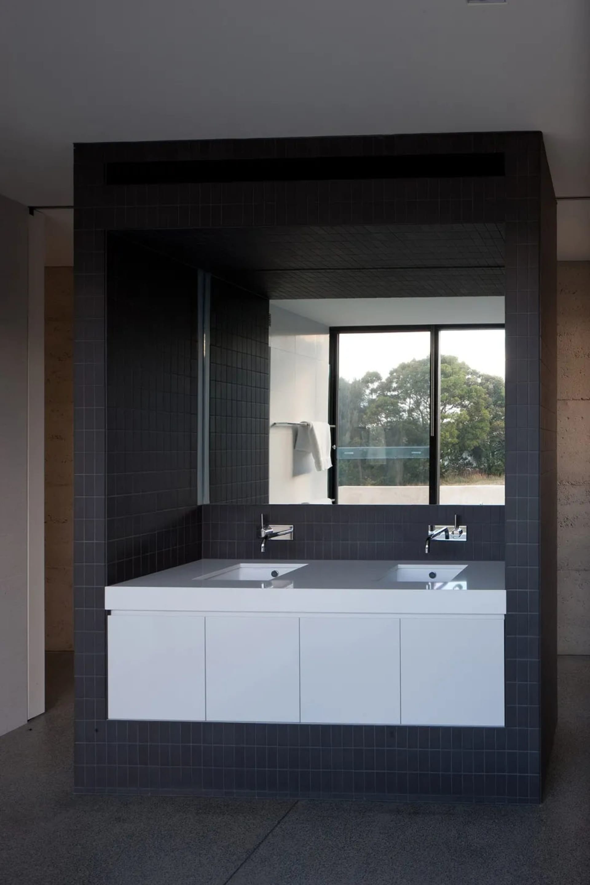 A modern residential bathroom with dark tiled walls, glass shower doors, and a concrete countertop.