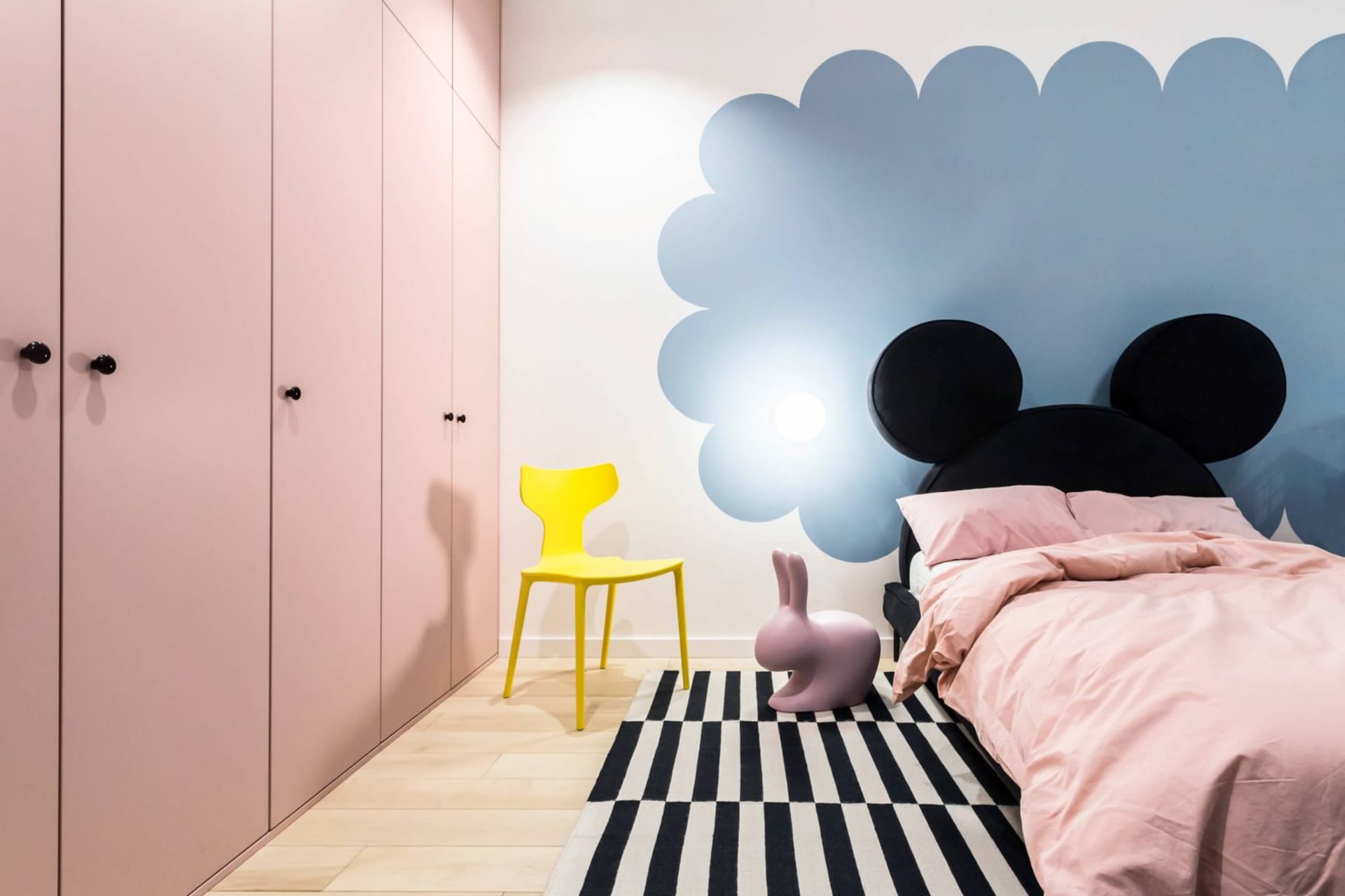 Minimalist bedroom featuring a pink bed with black Mickey Mouse ears headboard, a yellow chair, and a black and white striped