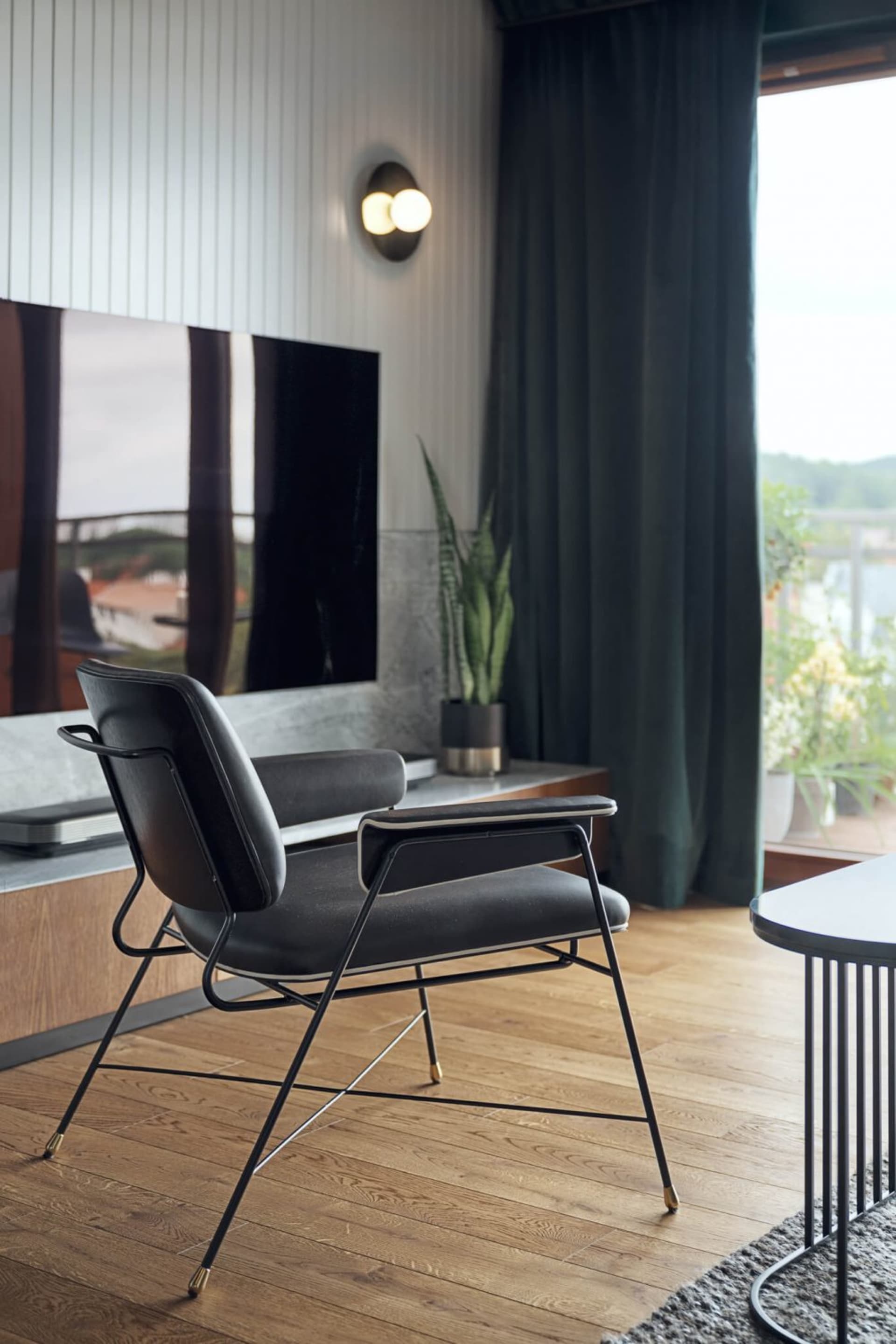 A modern minimalist living room featuring a wooden floor, black leather armchair, and a large window with green curtains.