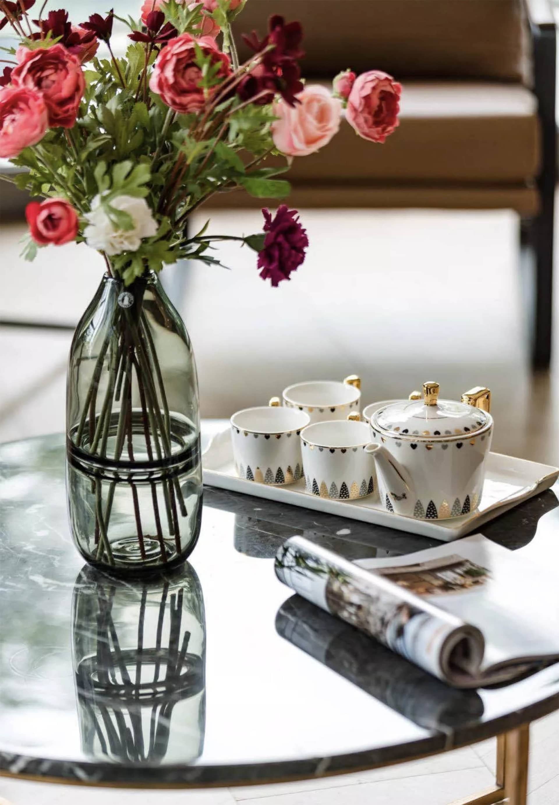 A modern interior featuring a glass coffee table with a vase of vibrant flowers, a teapot and cups, and a rolled-up magazine.