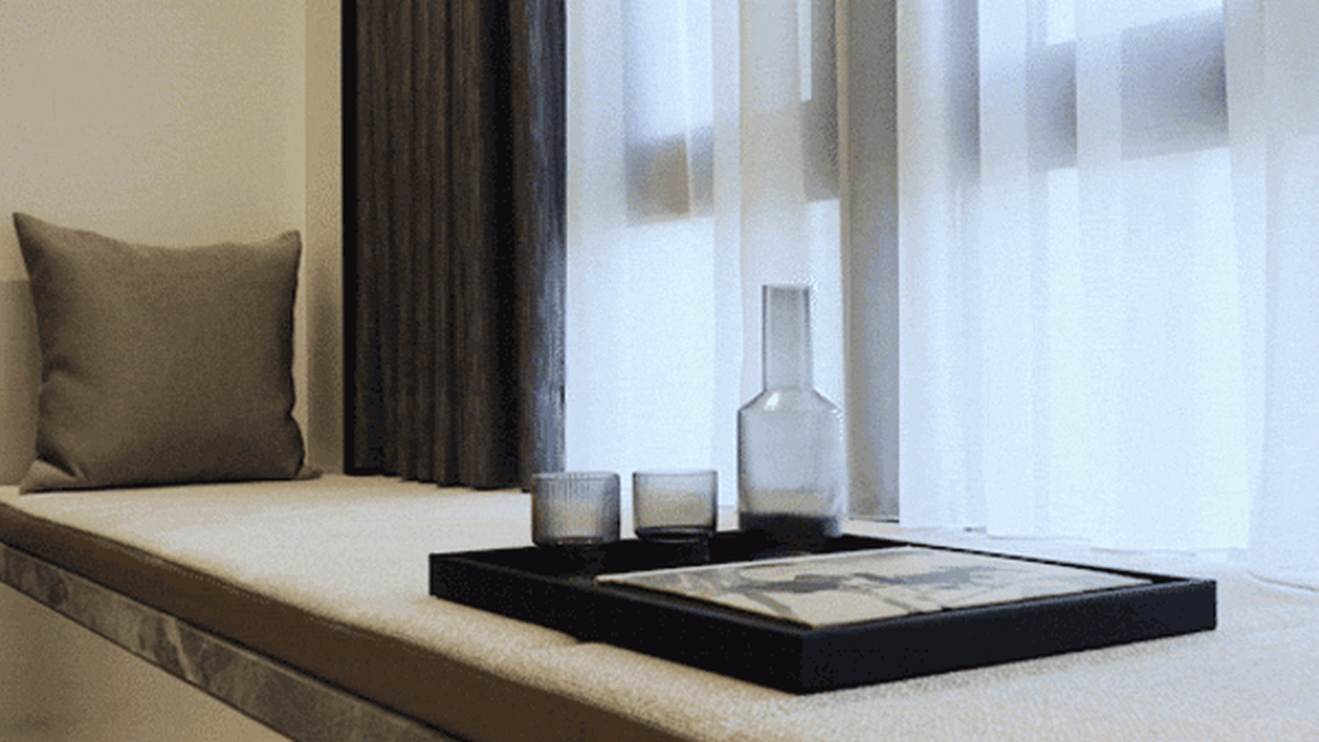 Modern minimalist bedroom featuring gray curtains, white bedding, and a black tray with a carafe and glasses on a cushioned b