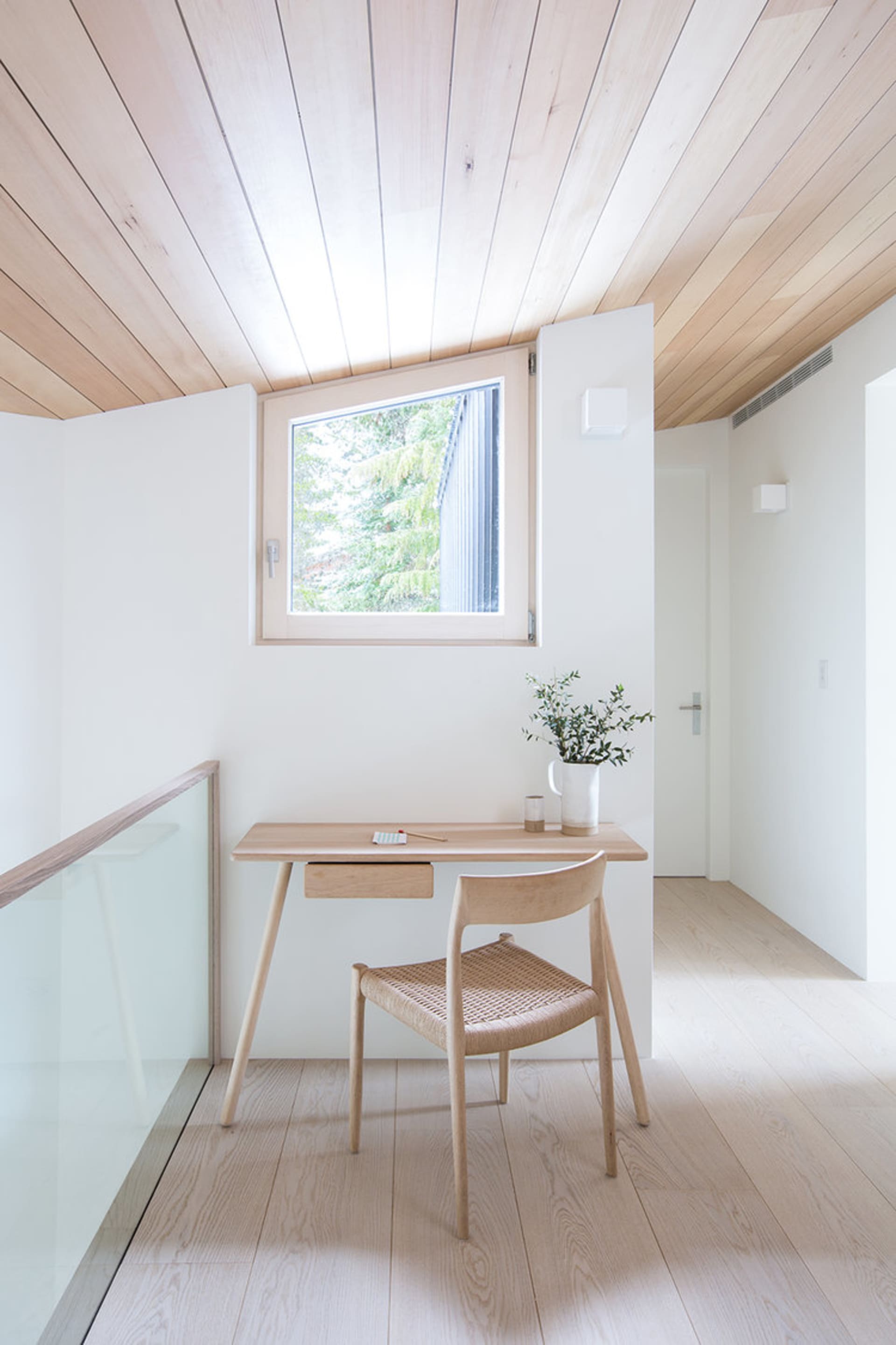 A minimalist home office with a wooden desk and chair, featuring a small window and natural lighting.