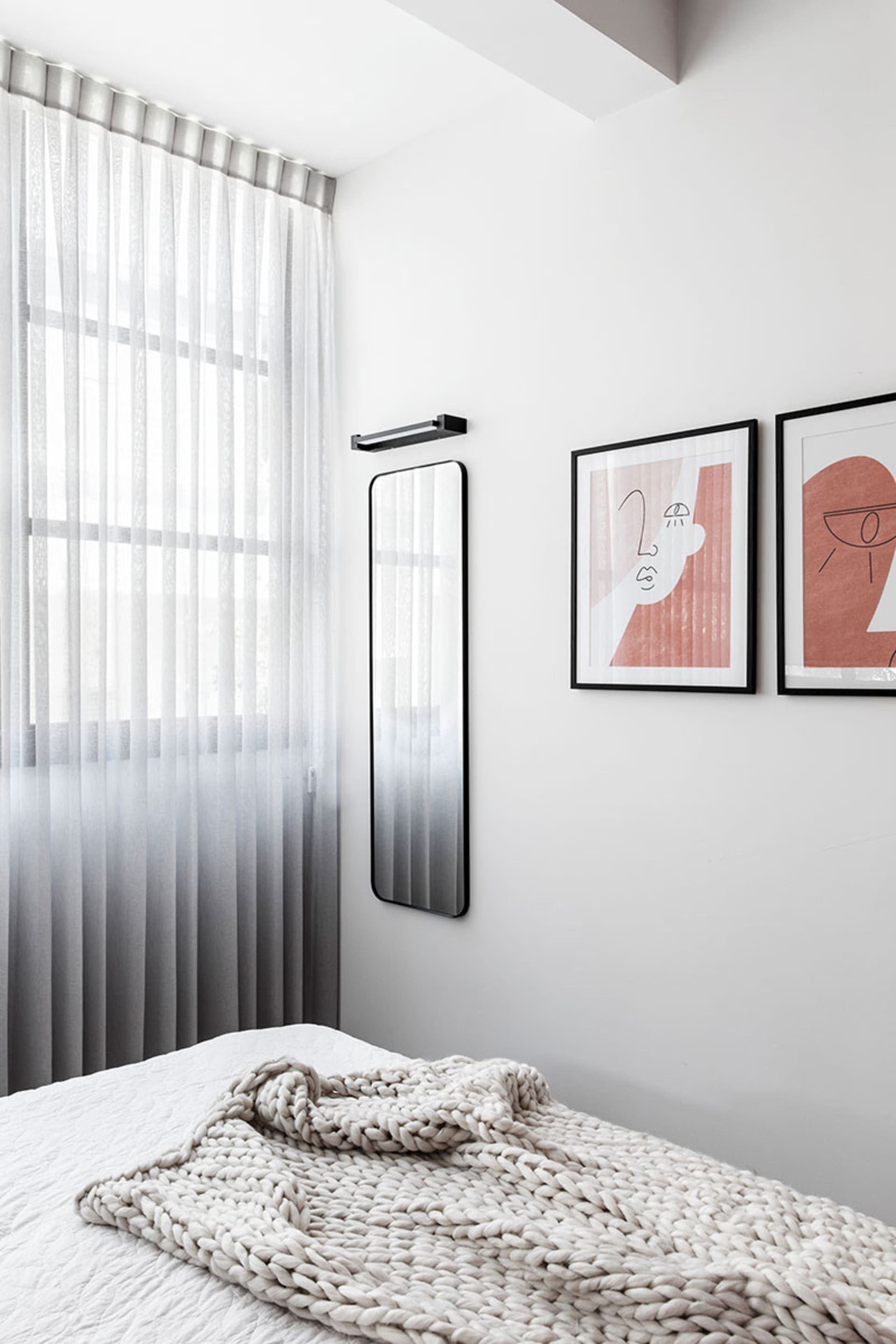 A minimalist bedroom featuring a white bed with a textured blanket, framed artworks, and sheer curtains allowing natural ligh