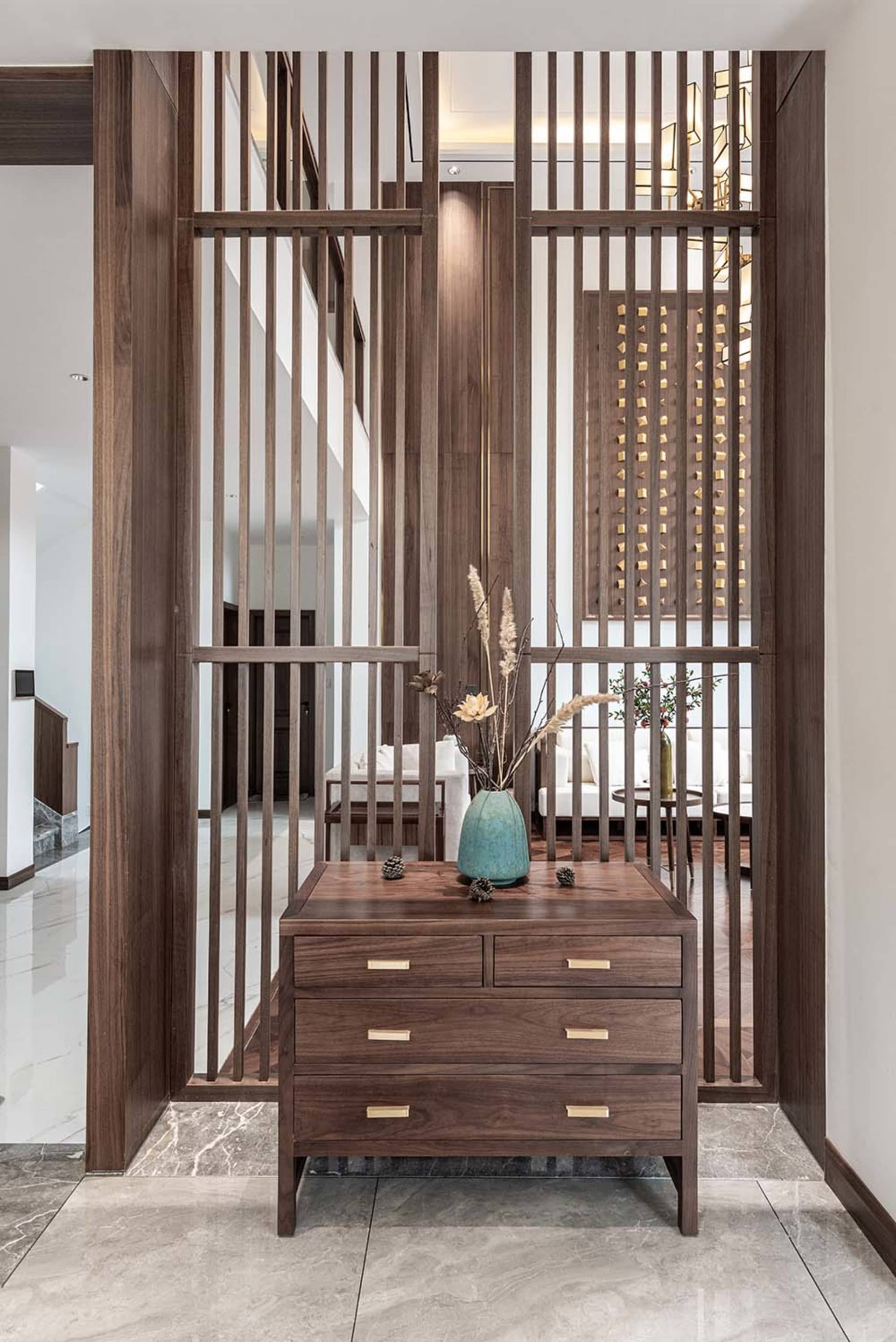 Modern entrance foyer with wooden slat partition, marble floor tiles, and decorative wooden chest with vase and dried plants.