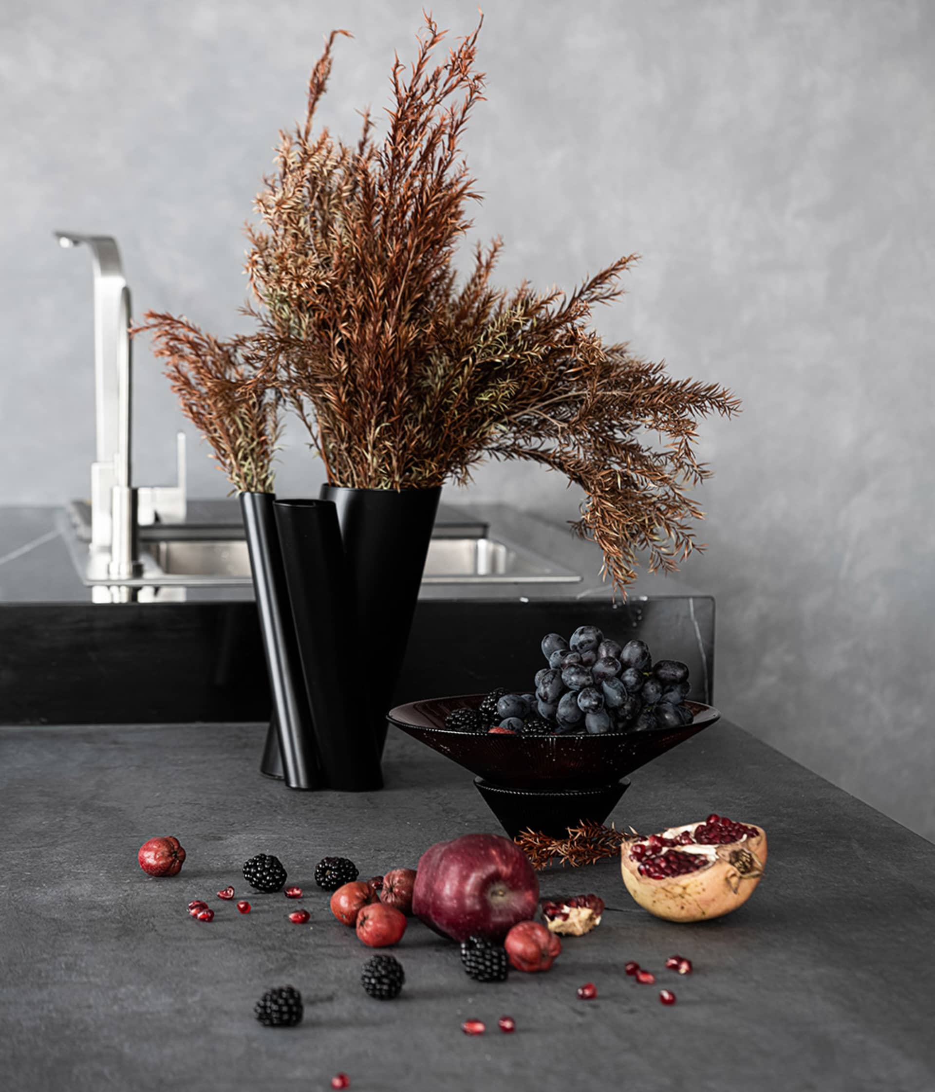 A modern kitchen interior with concrete countertops, a stainless steel faucet, a bowl of grapes and berries, and a vase with