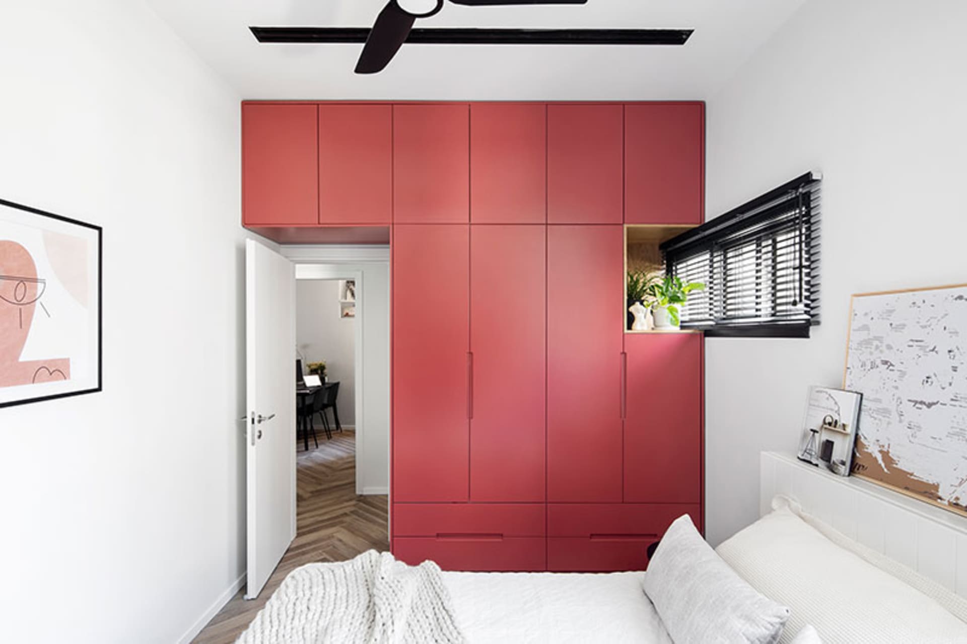 A modern minimalist bedroom featuring a large red cabinet, white bed, and white walls, with a view into a hallway with herrin