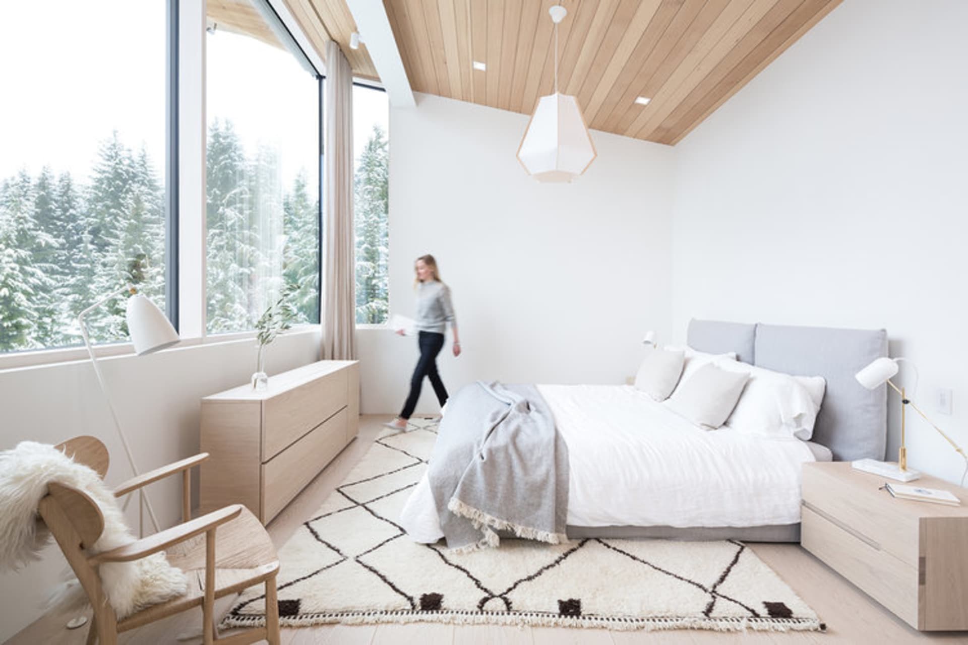 A modern minimalist bedroom with white walls, wooden furniture, and a large window overlooking snowy trees.