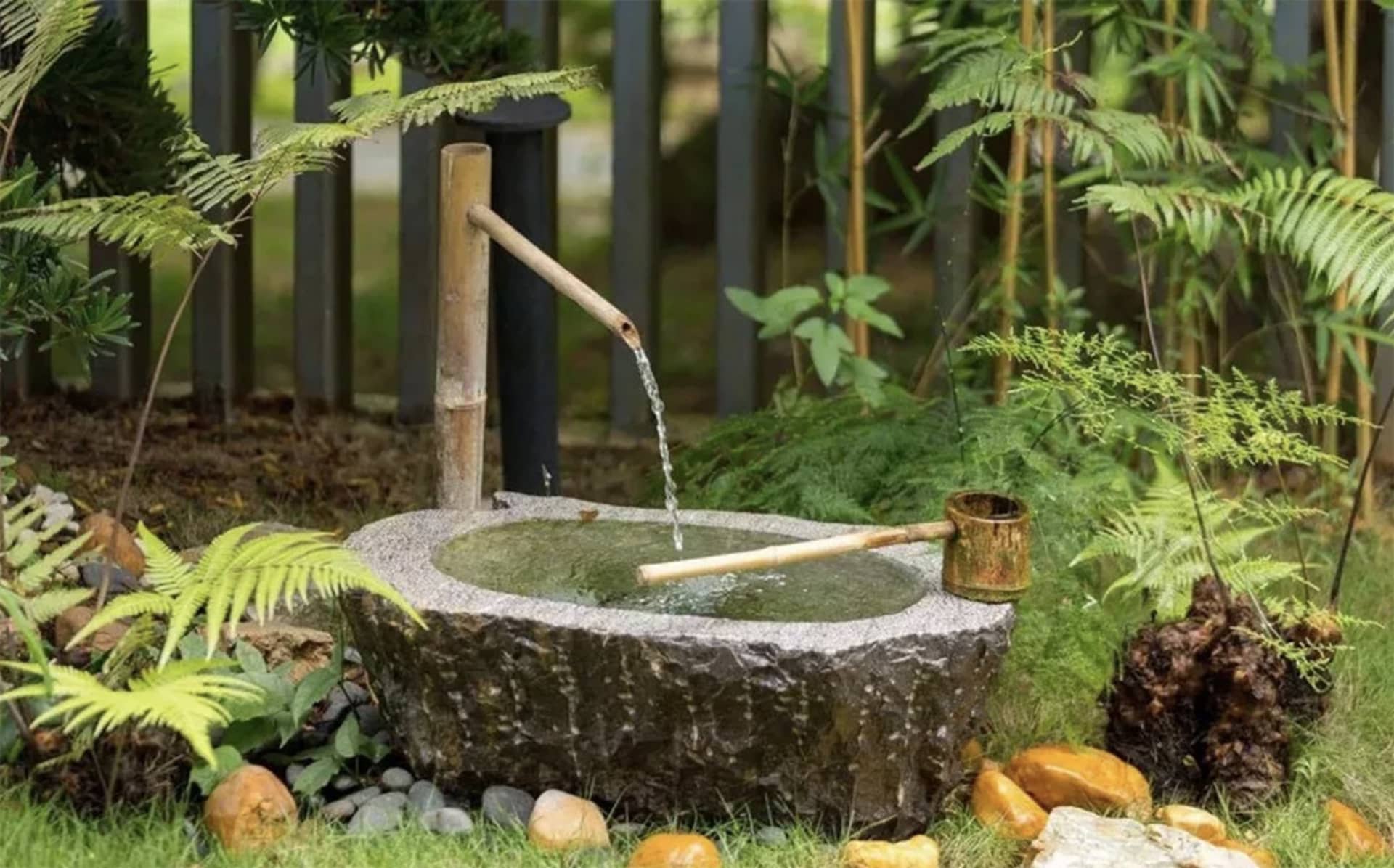 rustic stone water feature with a bamboo spout surrounded by green plants and rocks.