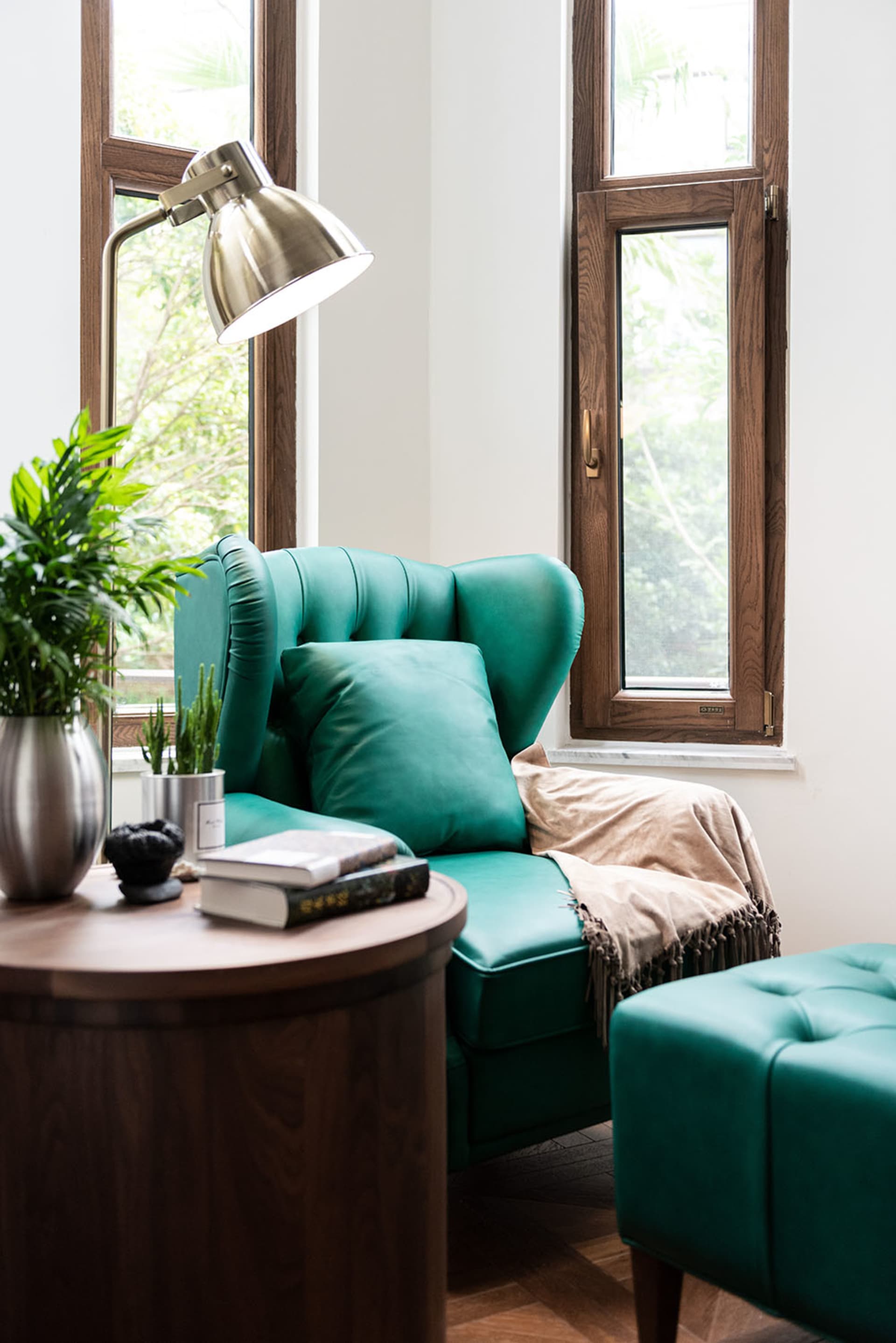A modern green upholstered armchair with a bright lamp, books, and a vase of plants in a cozy living room near a window.