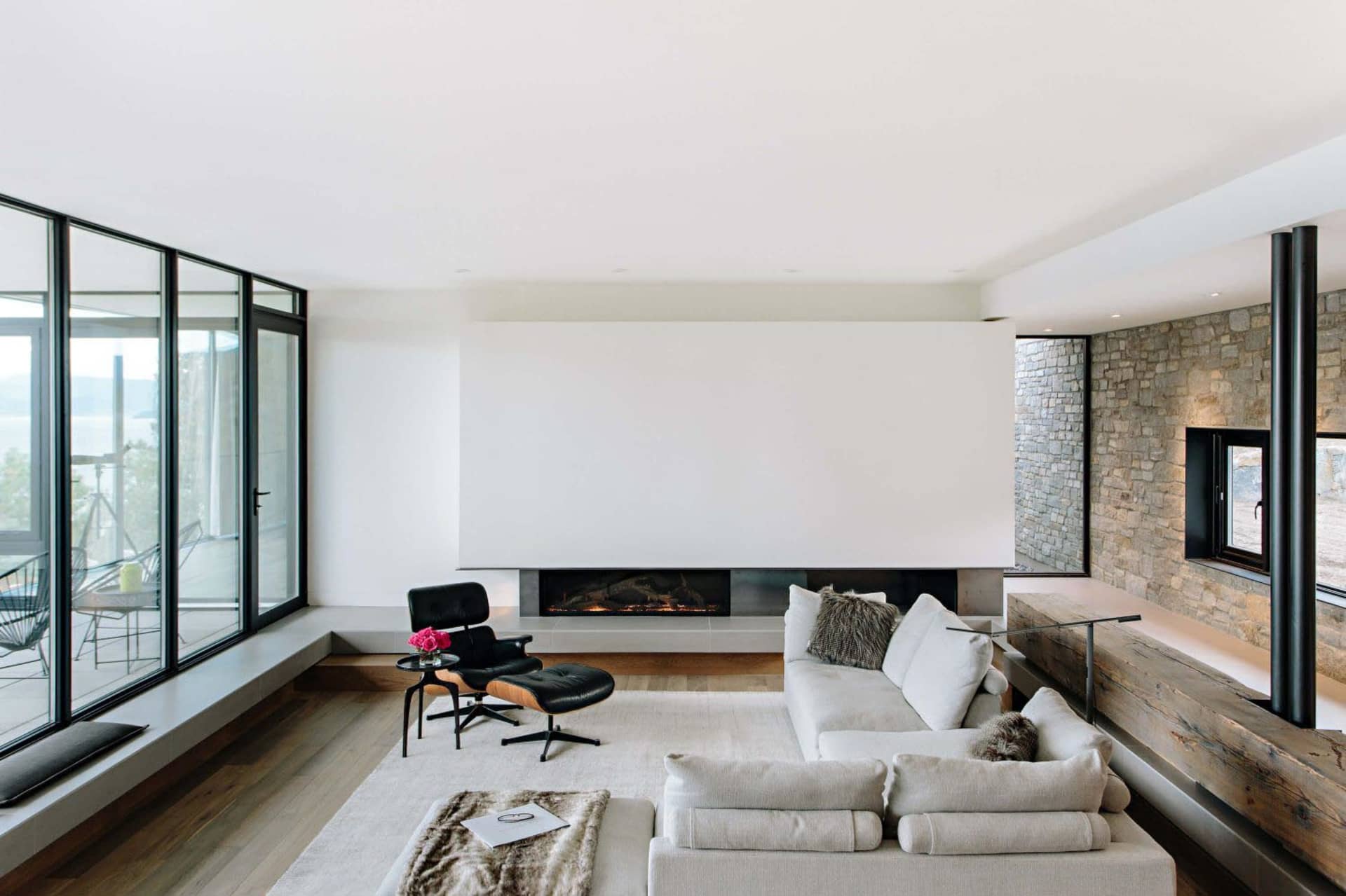 Modern living room with a fireplace, a white sectional sofa, and a black armchair with a footstool. The room features a stone