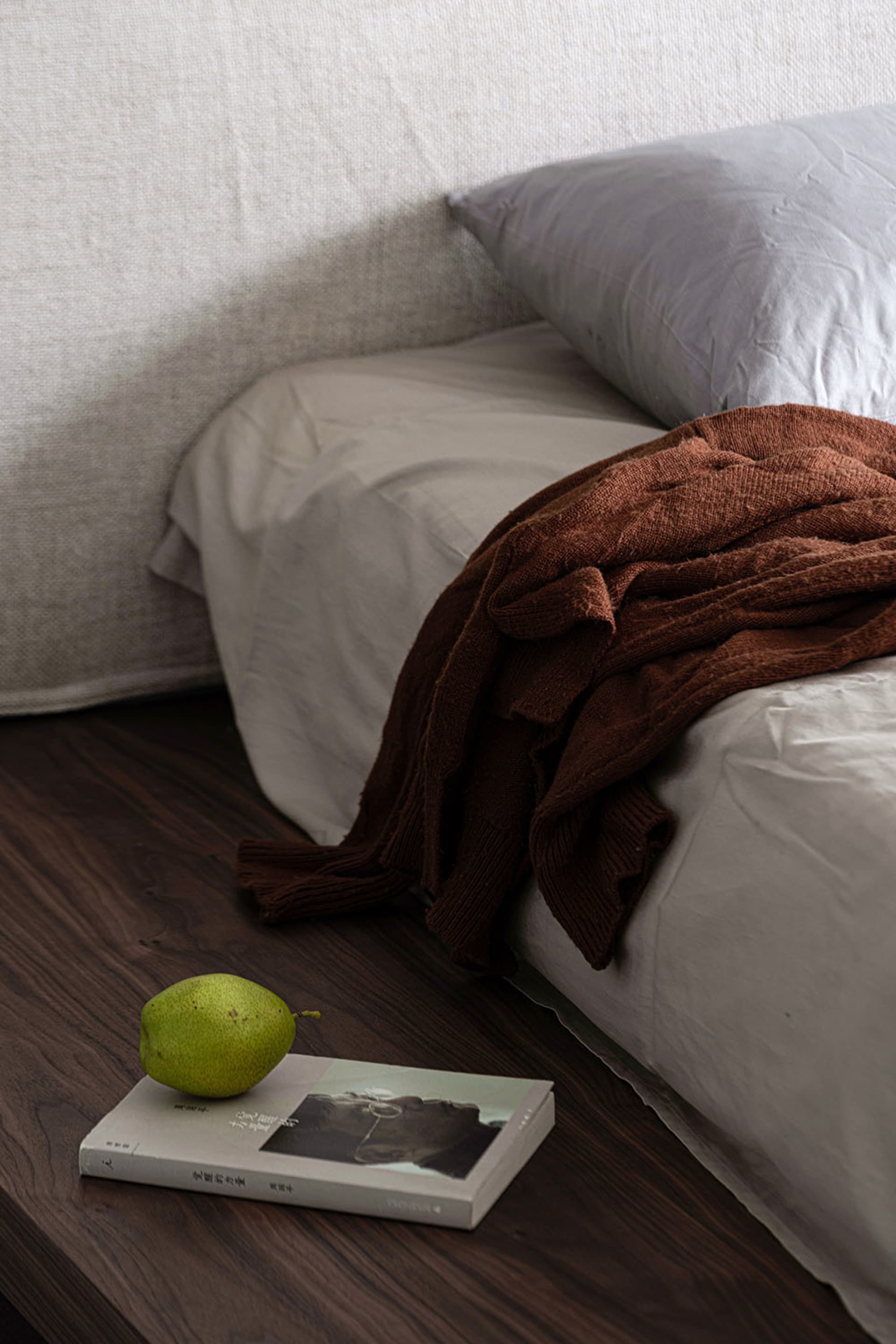A cozy minimalist bedroom with a bed covered in white linen and a brown knit sweater draped over it, a green pear resting on