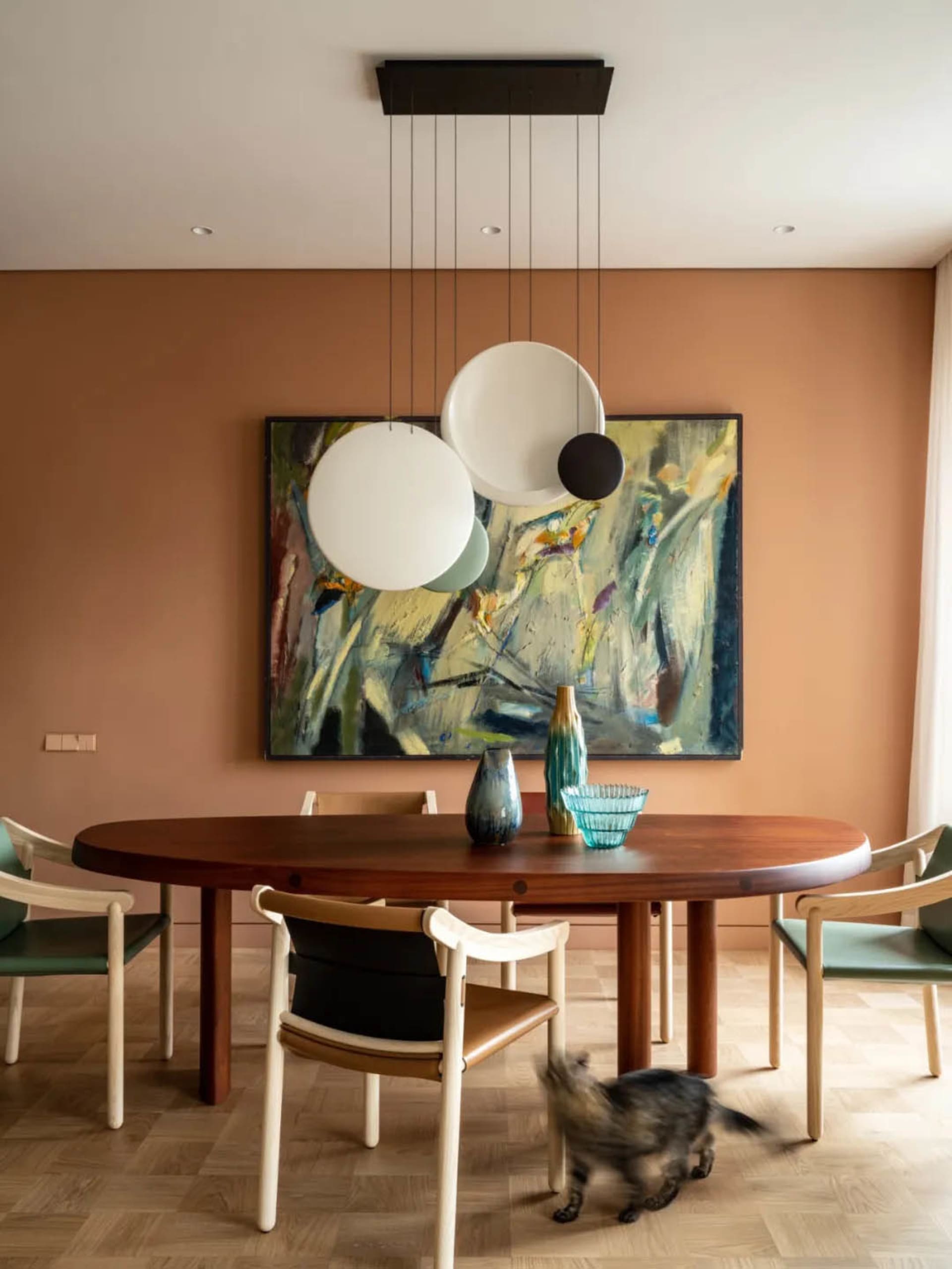 A modern dining room featuring a wooden table surrounded by chairs, a hanging light fixture, and a colorful painting on the w