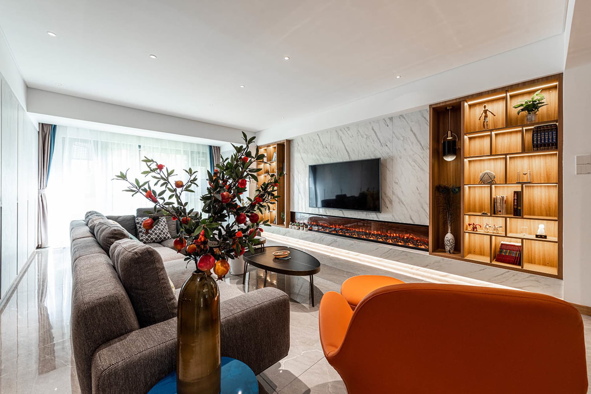 A modern living room featuring a grey sofa, orange chairs, pomegranate plant, glass vase, and wooden shelves with lighting.