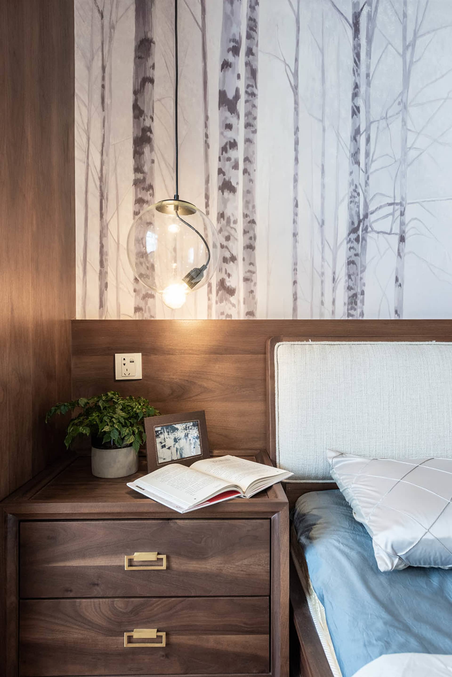 A modern minimalist bedroom featuring a wooden bed frame and nightstand with a potted plant, an open book, and a spherical pe