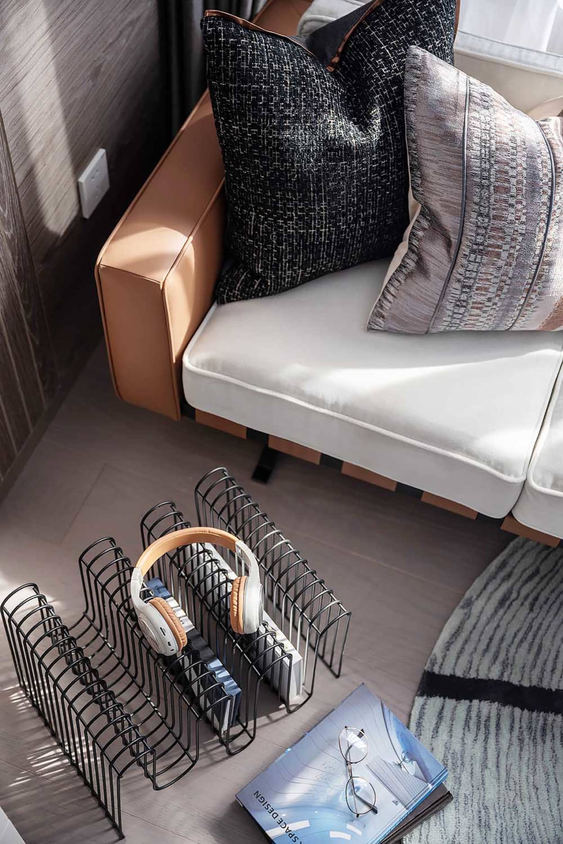 A modern living room with a sleek, minimalist sofa, textured pillows, and a wire magazine rack holding headphones and books.