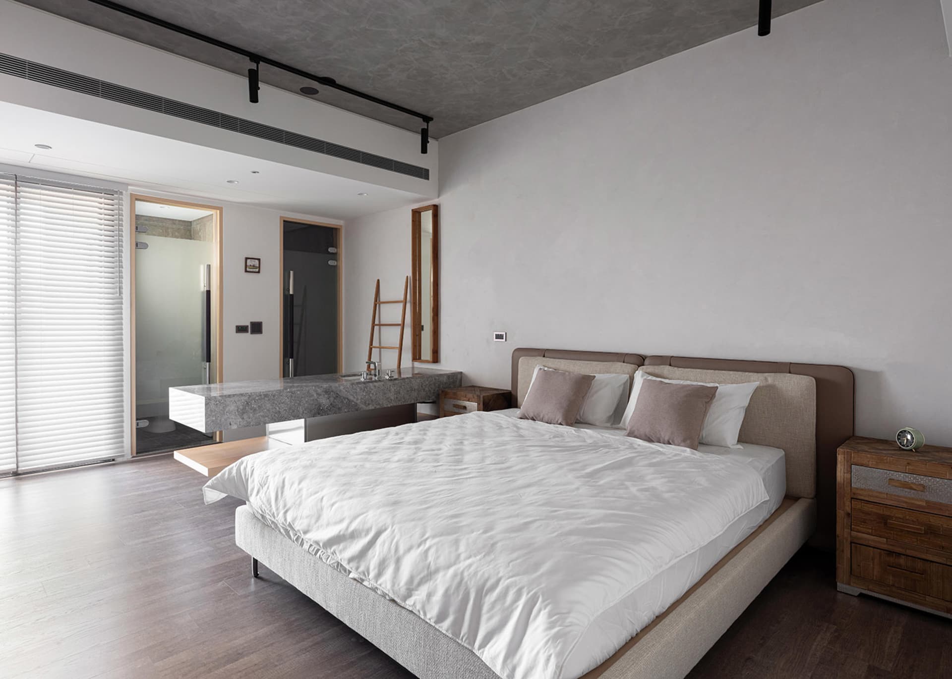 A minimalist bedroom featuring a bed with a concrete headboard and wooden furniture, in a modern industrial style.
