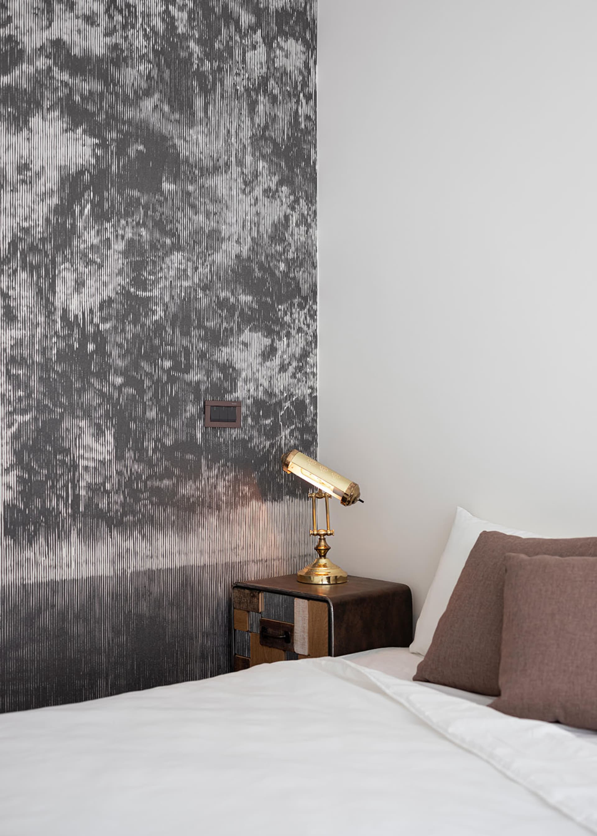 Minimalist hotel room featuring a textured wallpaper wall and a brass table lamp on a wooden bedside table.