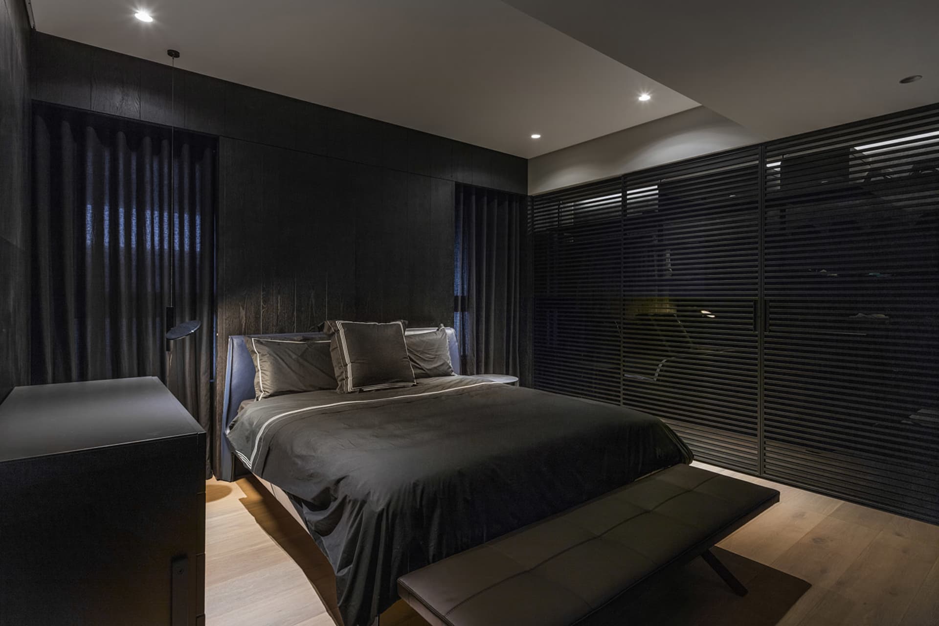 Minimalist bedroom featuring a wood bed with black bedding, a bench, and dark curtains.